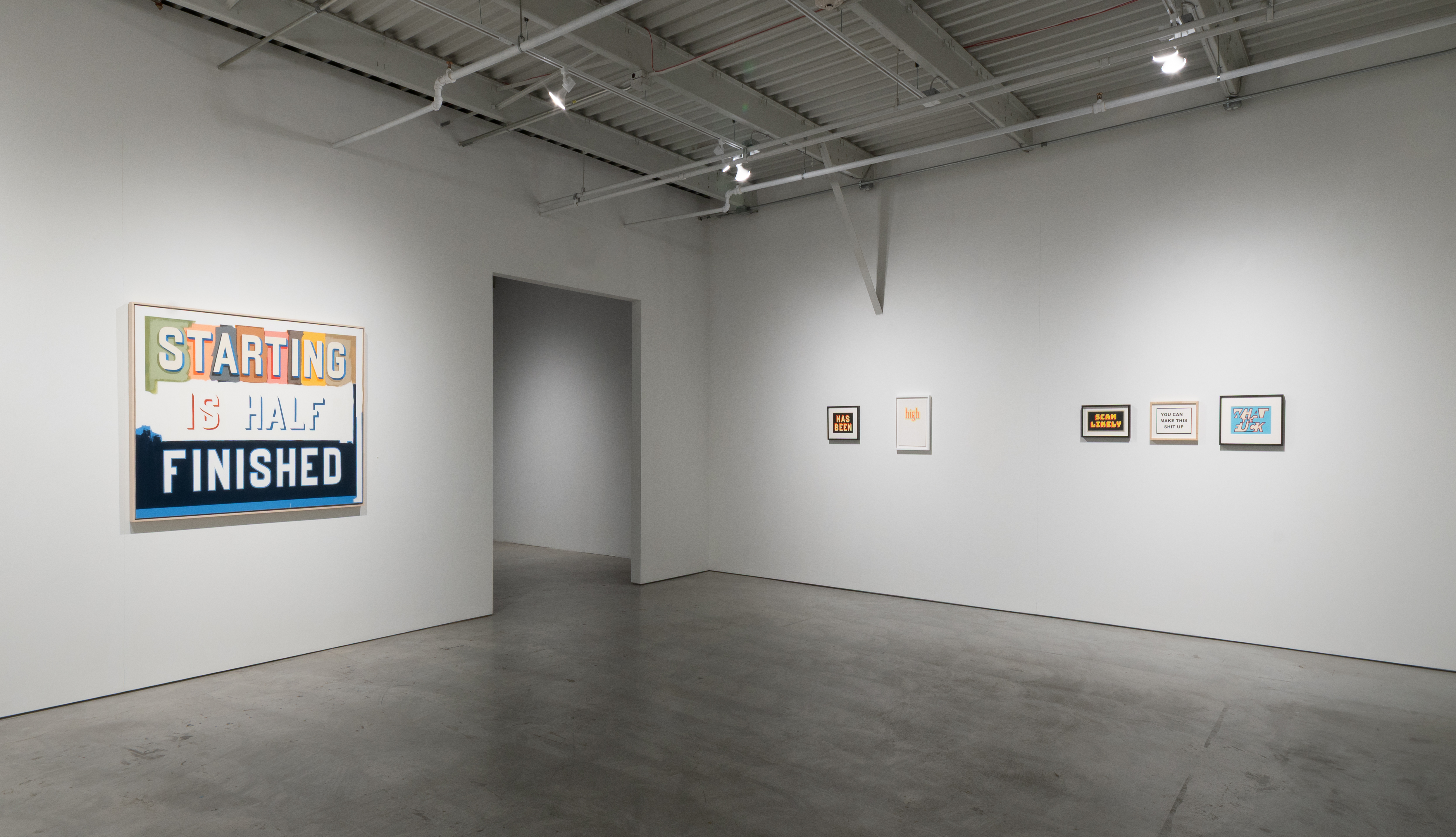 A gallery interior with white walls and industrial ceiling displays a large colorful sign reading "STARTING IS HALF FINISHED" on the left wall, alongside several smaller framed works with text-based imagery arranged on the right wall.