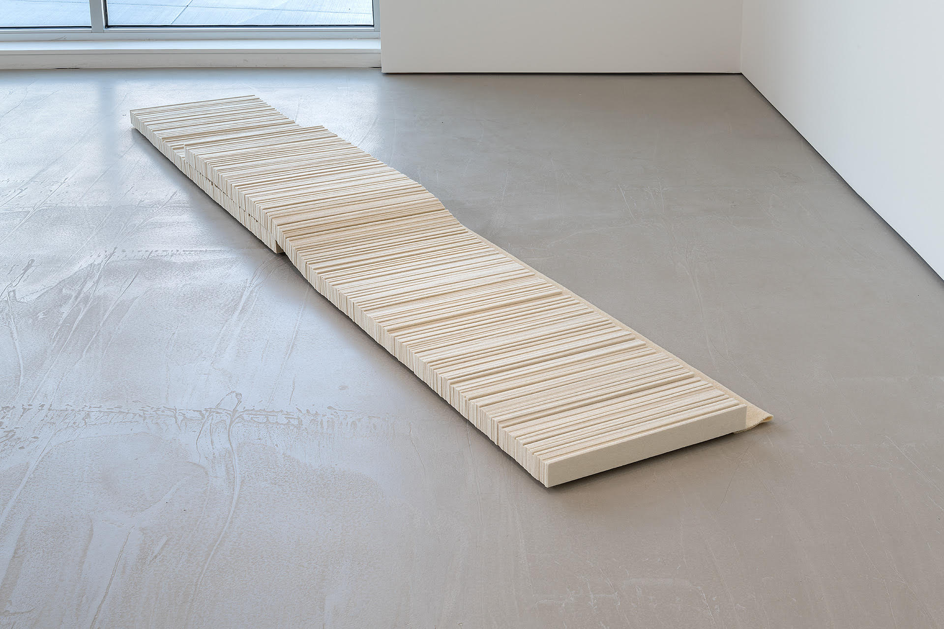 A long, narrow wooden slab with horizontal striations lies diagonally across a concrete floor in a minimalist gallery space. The natural wood grain contrasts with the smooth, neutral-toned surfaces surrounding it, creating a simple geometric composition that emphasizes material and shadow.