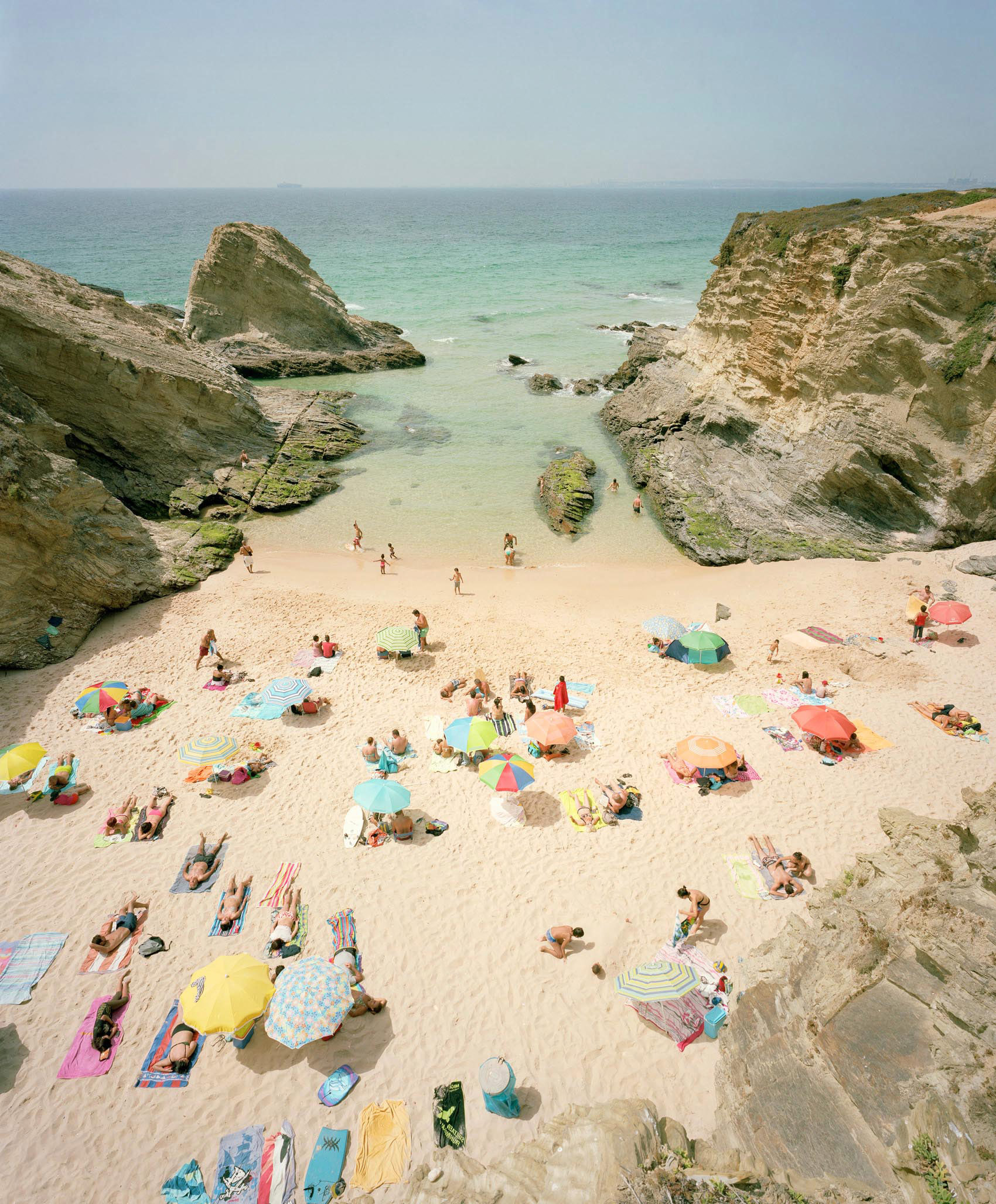 Aerial view of a crowded beach nestled between dramatic limestone cliffs, with colorful umbrellas and sunbathers scattered across golden sand and shallow turquoise waters, rocky outcrops visible in the cove below.