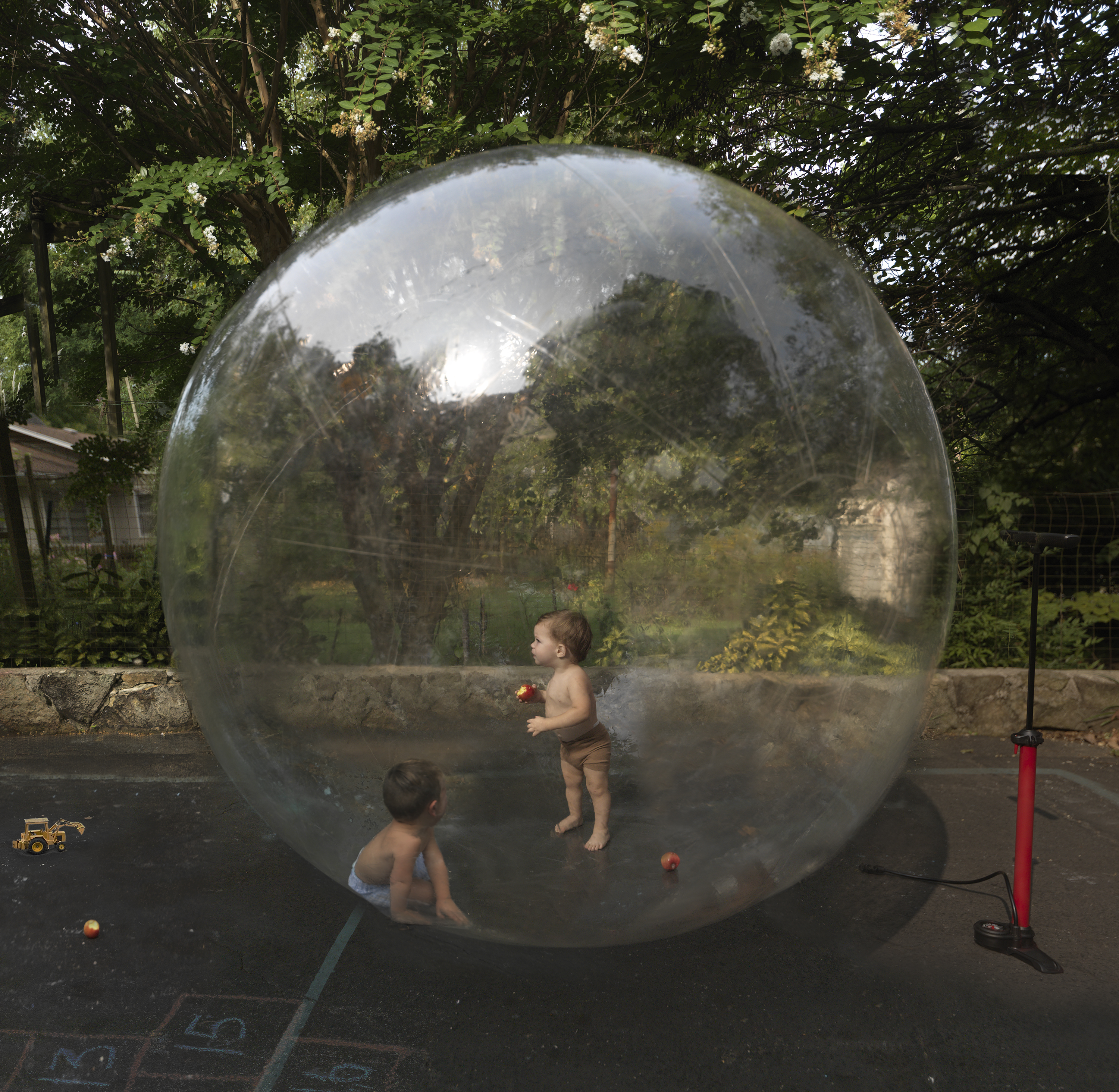 Two young children play inside a large transparent bubble in a residential driveway, surrounded by green foliage and trees. The spherical installation reflects and distorts the landscape, creating a luminous dome that contrasts with the dark asphalt ground below.