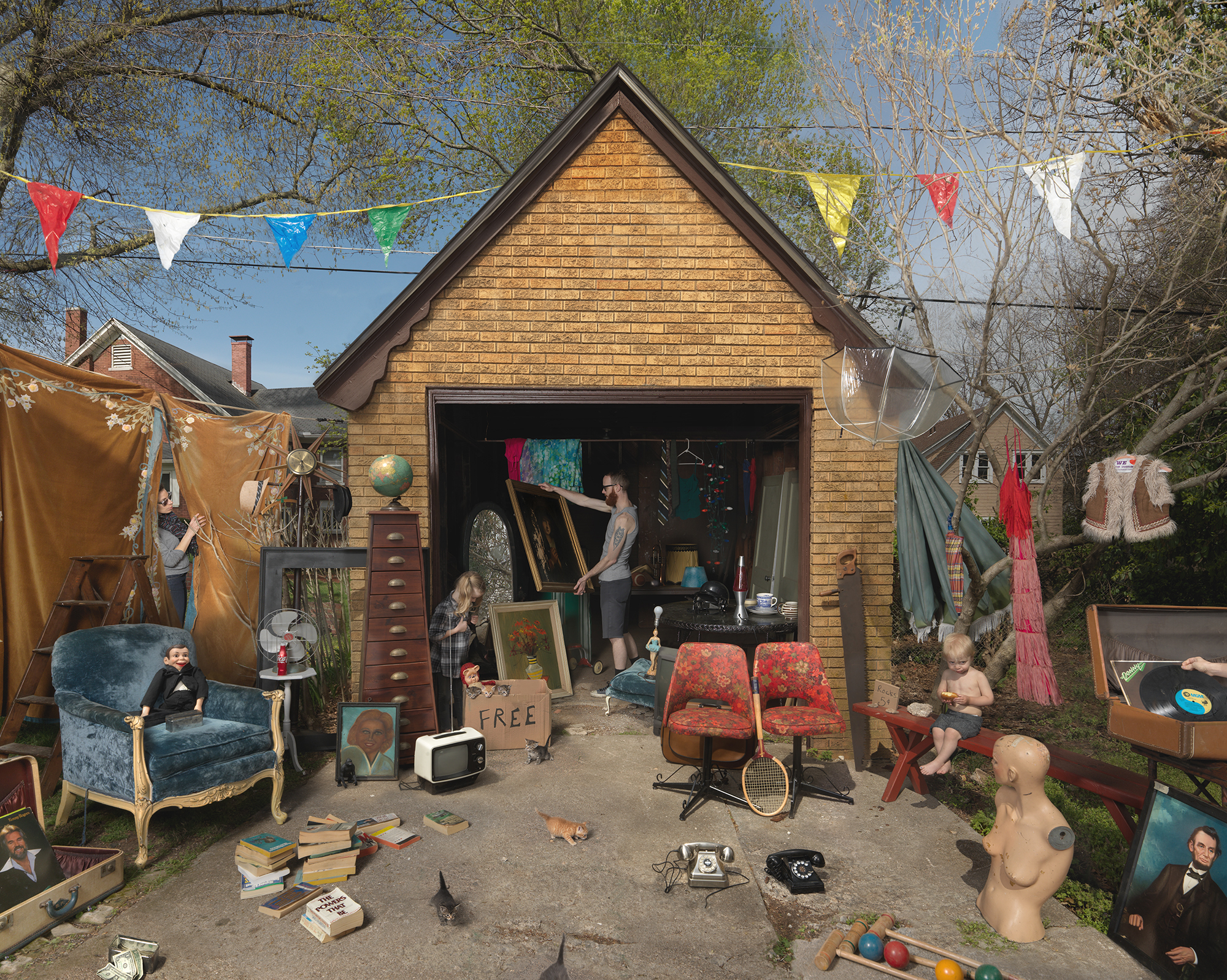 A suburban garage transformed into a bustling yard sale scene populated with mannequins and children among vintage furniture, artwork, textiles, and household goods. Colorful bunting flags string overhead while clothes hang from lines, creating a theatrical tableau of organized chaos and commerce.