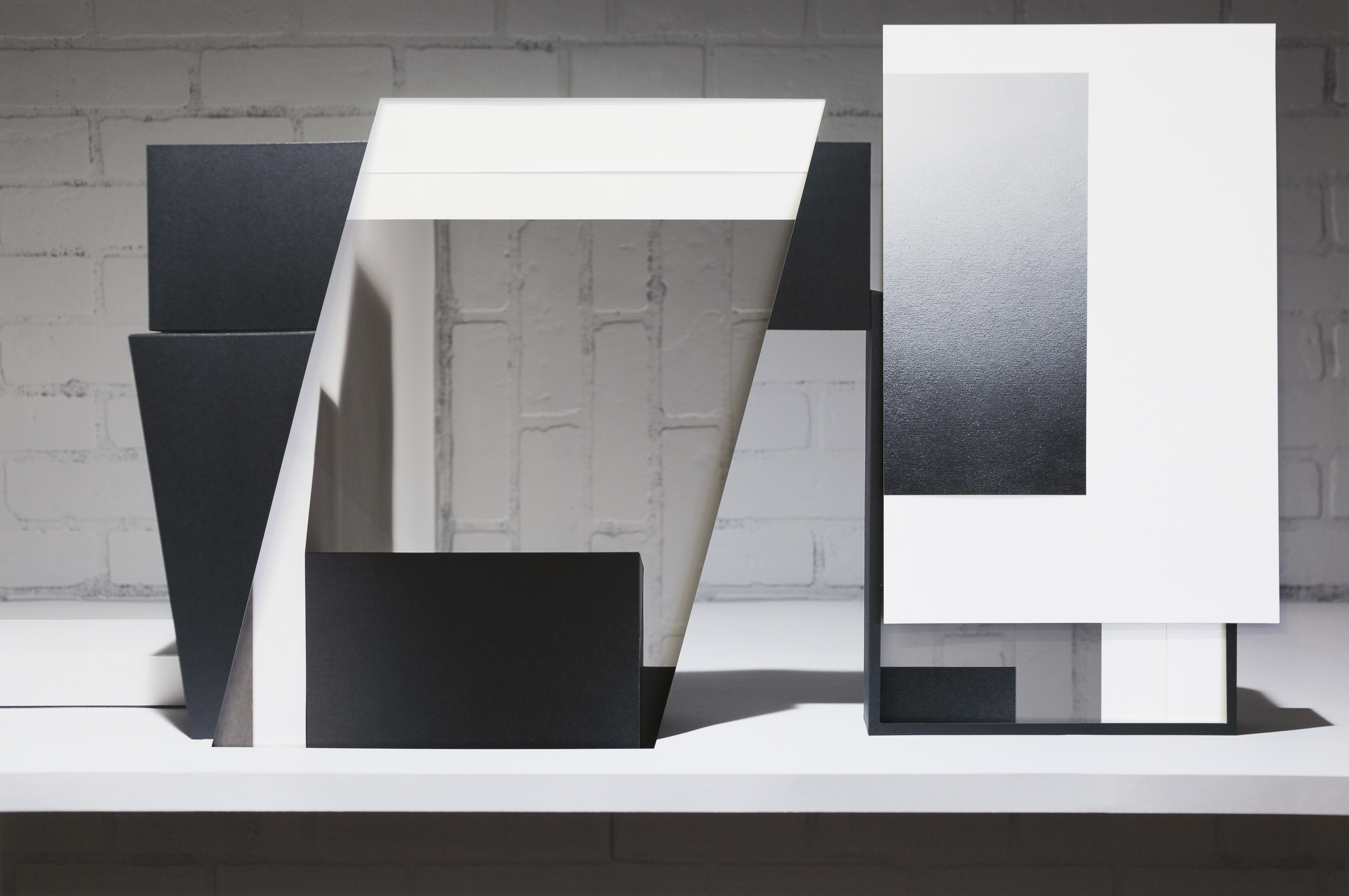 Abstract composition of collaged and layered white and black rectangular forms arranged on a white platform against a weathered brick wall, creating geometric planes that overlap and cast subtle shadows, with a minimalist aesthetic emphasizing contrasting values and spatial depth.