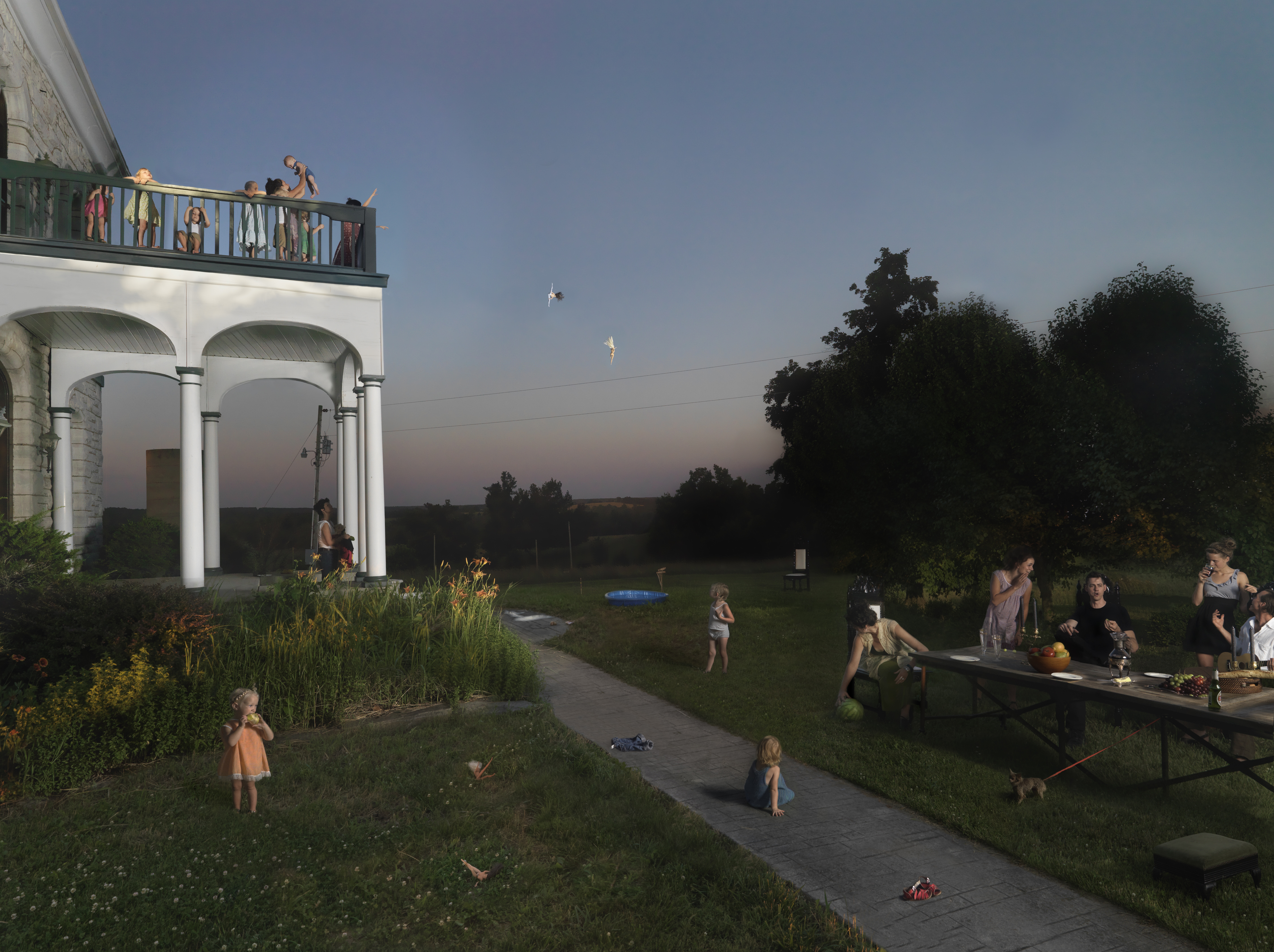 A domestic scene rendered in soft twilight tones shows families gathered in a sprawling lawn beneath a two-story white classical structure with arched colonnades. Children play across the grass and pathways while adults socialize on the right; figures lean over a railing on the upper balcony. The composition captures an everyday moment of leisure with warm, naturalistic lighting against a dusky sky.