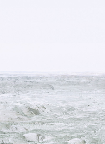 Ethereal landscape photograph of an expansive, barren terrain with undulating hills rendered in soft whites, grays, and pale greens beneath a hazy, overexposed sky. The composition creates an abstracted, almost monochromatic vista that blurs the distinction between earth and atmosphere.