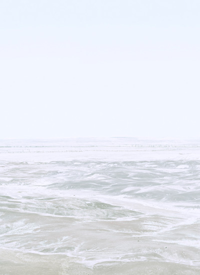 Soft, hazy seascape with gentle waves and foam washing onto a pale sandy beach beneath an overcast sky, rendered in muted tones of white, gray, and soft green in a minimalist composition.