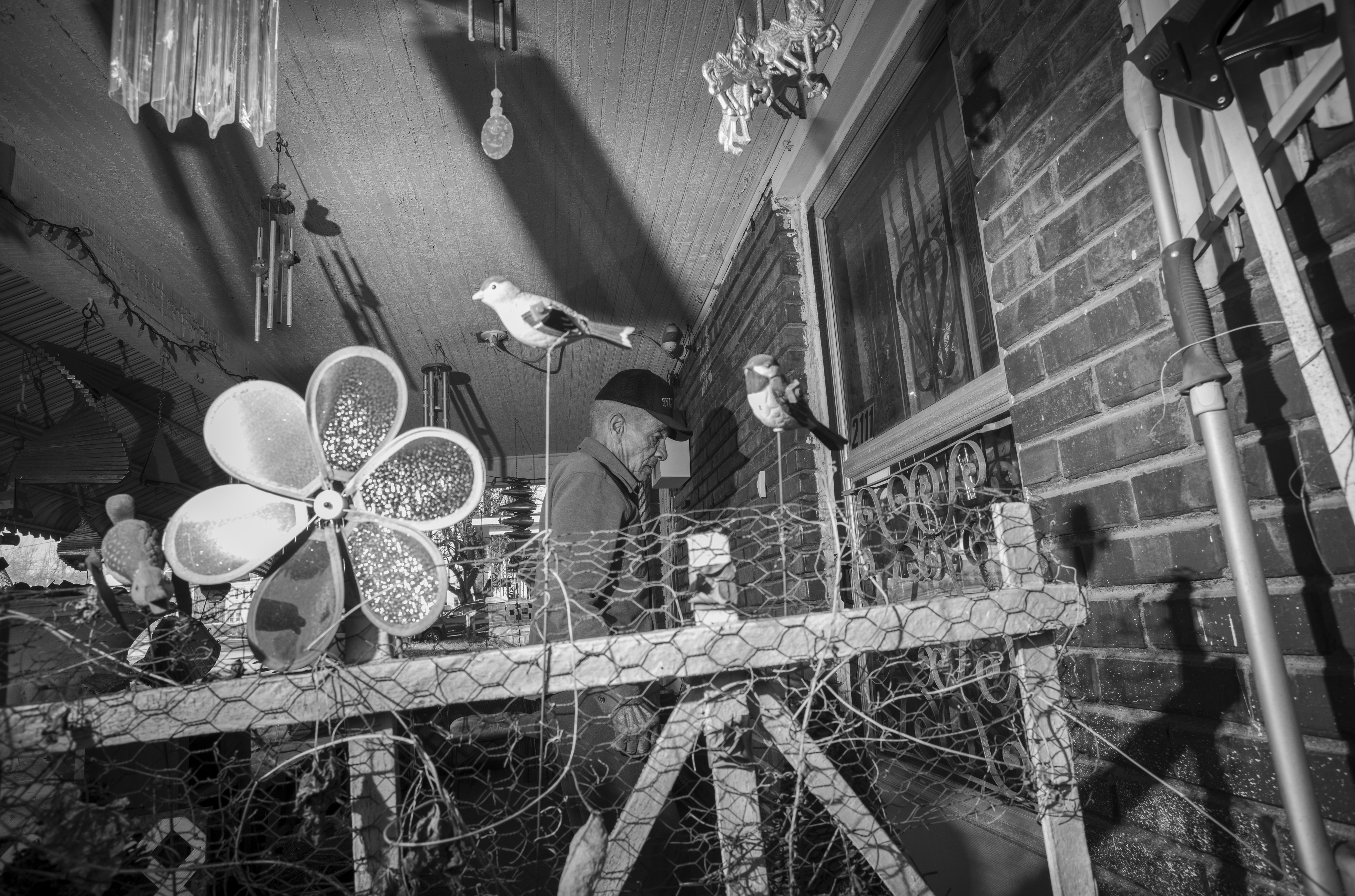 Black and white photograph of an older man in a cap standing in a narrow enclosed space with brick walls, surrounded by hanging wind chimes, decorative bird figurines, and potted plants with delicate metal flowers, creating a densely layered composition of domestic objects and ornaments.
