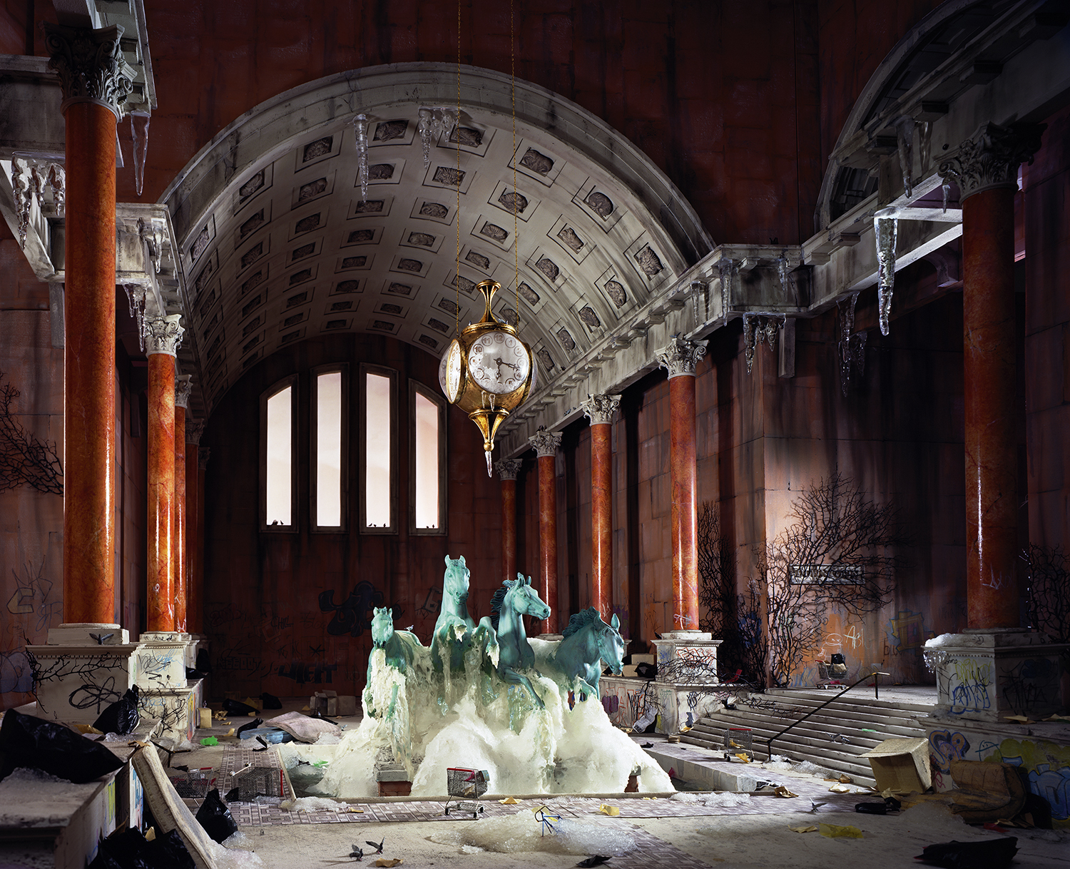 Abandoned interior of a grand neoclassical hall with soaring barrel-vaulted ceiling, ornate coffers, and massive red marble columns. A verdigris copper horse sculpture fountain sits centrally on the dusty floor beneath a hanging brass clock, while bare branches and debris scatter throughout the cavernous space lit by tall arched windows.