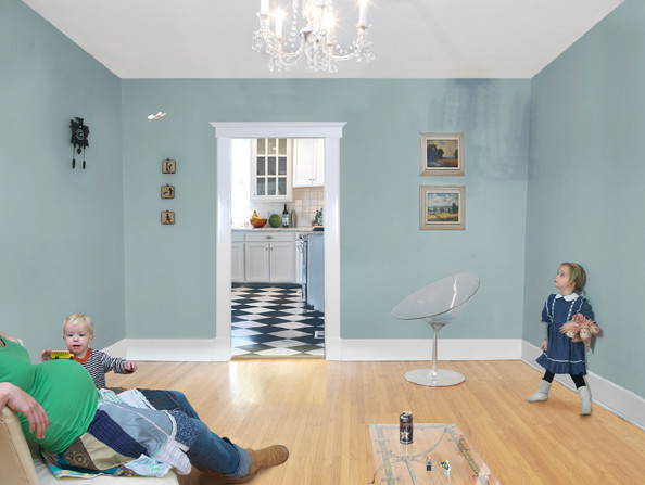 A light-filled domestic interior with pale blue-green walls features two children playing in a spacious room with blonde wood flooring. An adult in a green shirt reclines on the left while a young girl stands on the right; a modern white chair sits between them. Sunlight streams through a doorway in the center, revealing a black and white checkered kitchen floor beyond, with framed artwork and small decorative objects arranged on the walls.