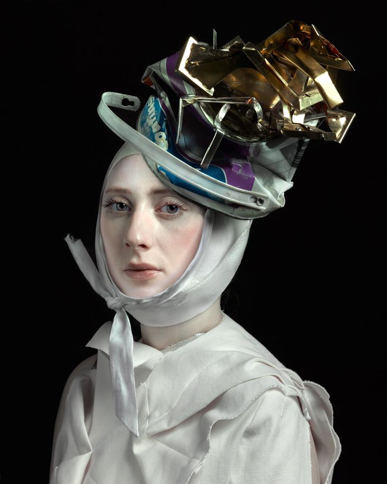 Portrait of a person wearing a white head covering and light garment, topped with an elaborate sculptural headdress constructed from metallic gold planes, purple fabric, and discarded shoe materials. Set against a black background, the composition blends classical portraiture with contemporary assemblage art.