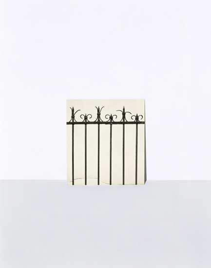 A minimalist composition featuring a decorative wrought iron fence with ornamental finials positioned against a pale cream-colored wall, casting sharp shadows on a light gray ground plane below.