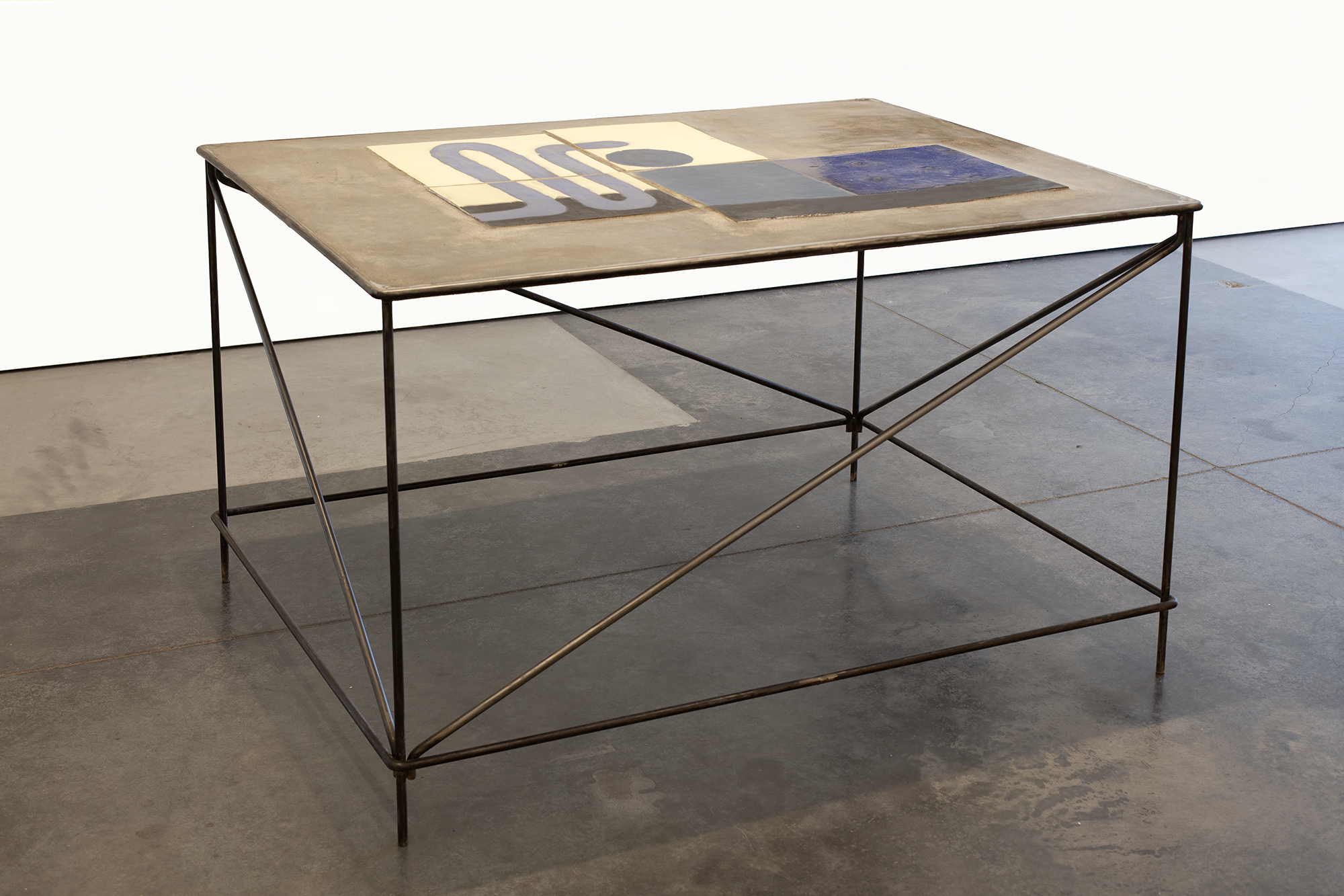 Minimalist table with a raw concrete top inlaid with geometric abstract shapes in cream, gray, and blue glazed ceramic. The steel frame features a geometric cross-bracing pattern on all sides, supporting a lower shelf and elevating the work on angled legs.