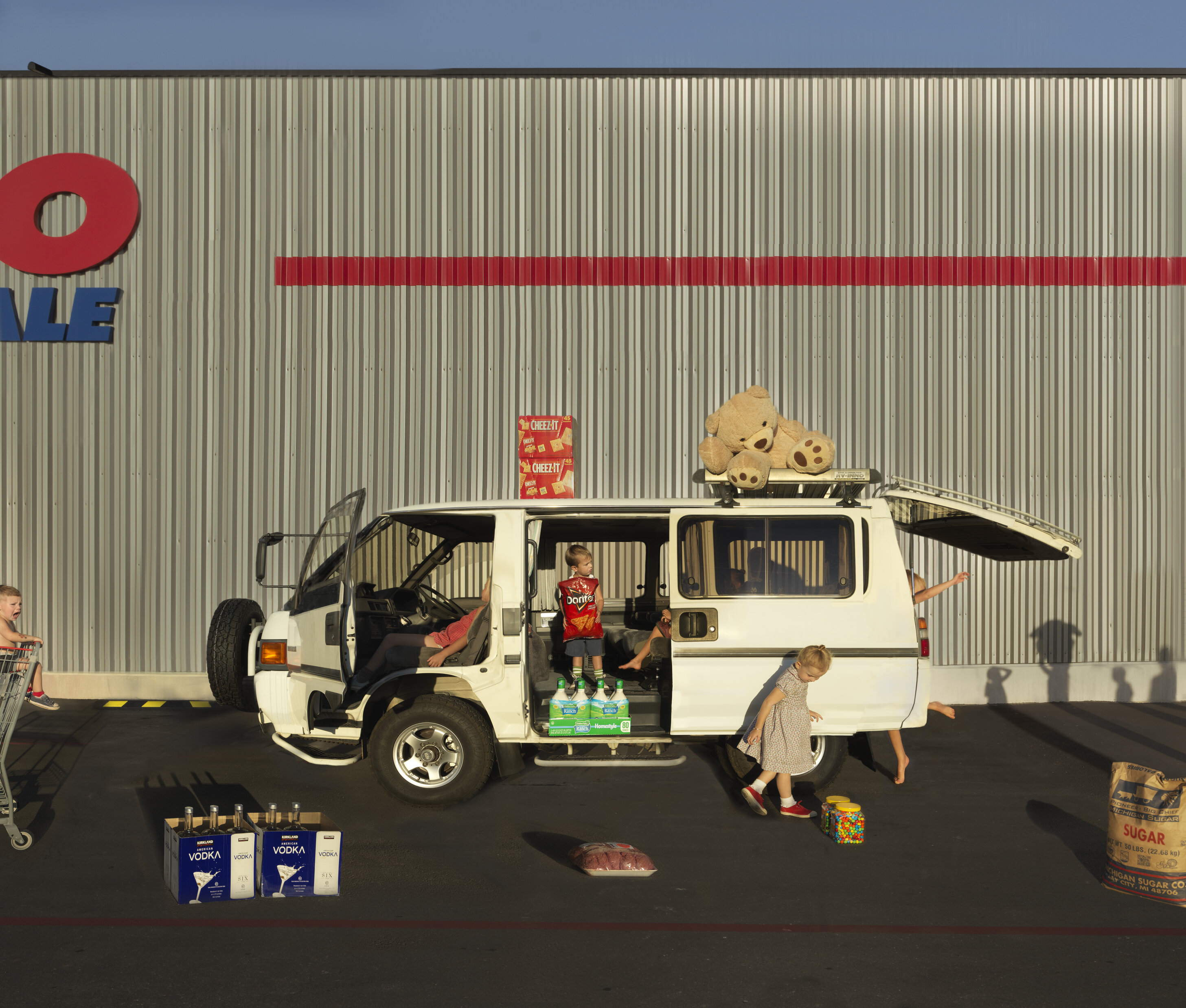 A white van with open doors is loaded with groceries and topped with teddy bears in front of a corrugated metal warehouse. Children play nearby among scattered consumer goods including vodka bottles, crackers, and sugar, arranged in a sprawling composition that extends across the ground plane against the industrial building's facade.