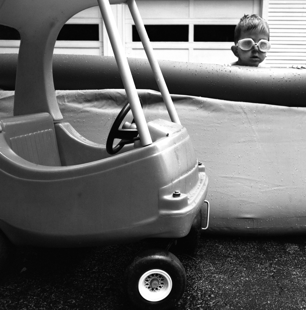 Black and white photograph of a young child wearing oversized goggles, partially visible inside a white toy airplane. The child peers out from the aircraft's interior while sitting among its molded plastic surfaces and control elements. In the foreground, a detailed wheel with a decorative hub is visible on the ground.