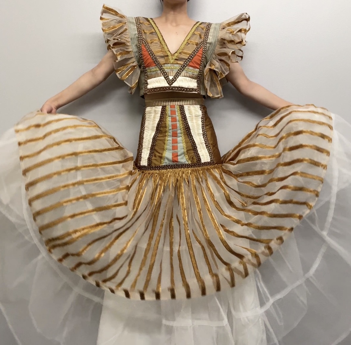 A figure wears an elaborate costume featuring a structured bodice with ornamental embroidery in earth tones, coral, and cream, paired with dramatically voluminous pleated skirt panels in cream and gold striped fabric. Ruffled cap sleeves and a full skirt create an exaggerated silhouette reminiscent of theatrical costume design.