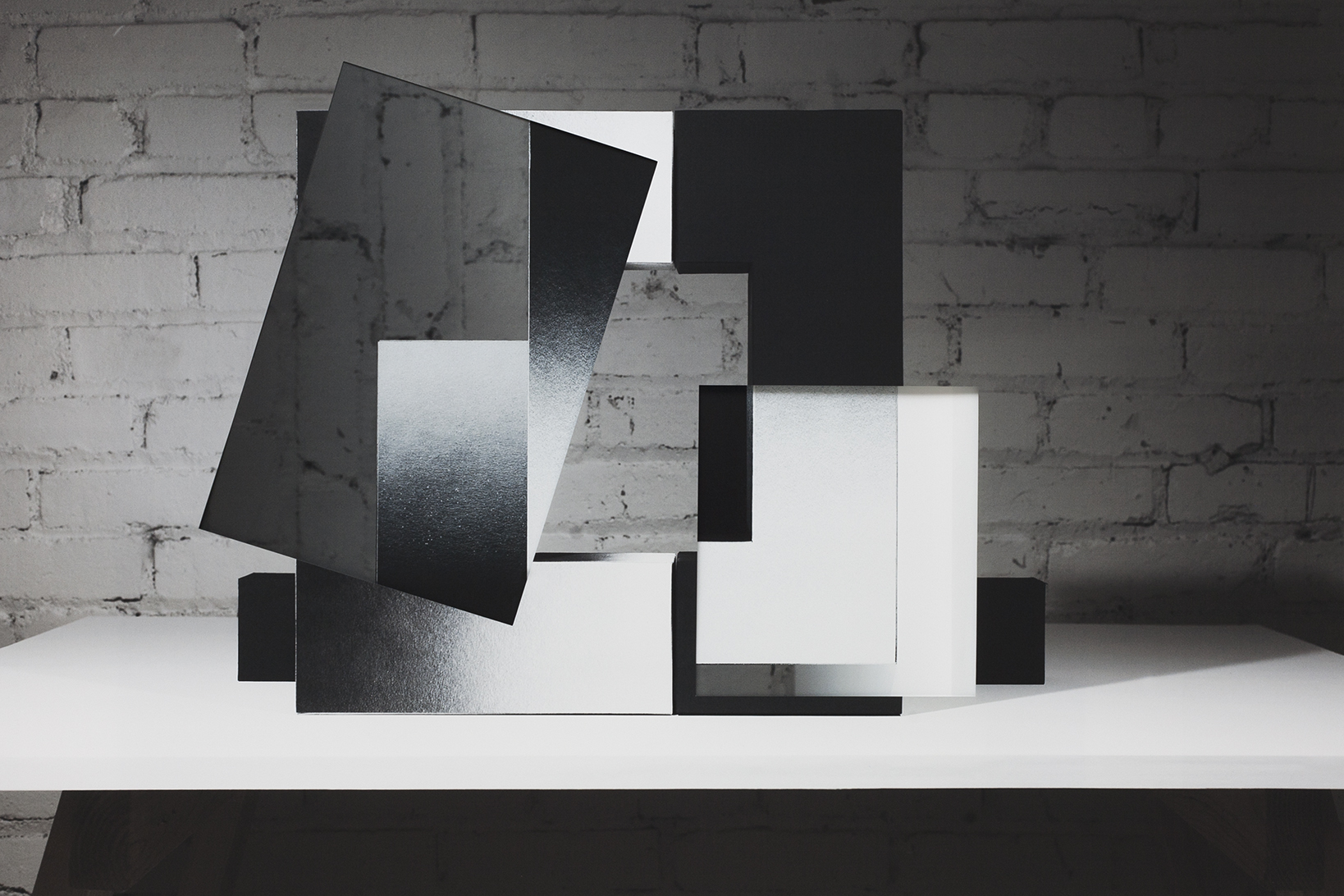 Abstract geometric composition made from layered black and white rectangular forms arranged in an interlocking, three-dimensional configuration. The work features torn and peeling paper elements creating varied surface textures, mounted on a white platform against a weathered brick wall.