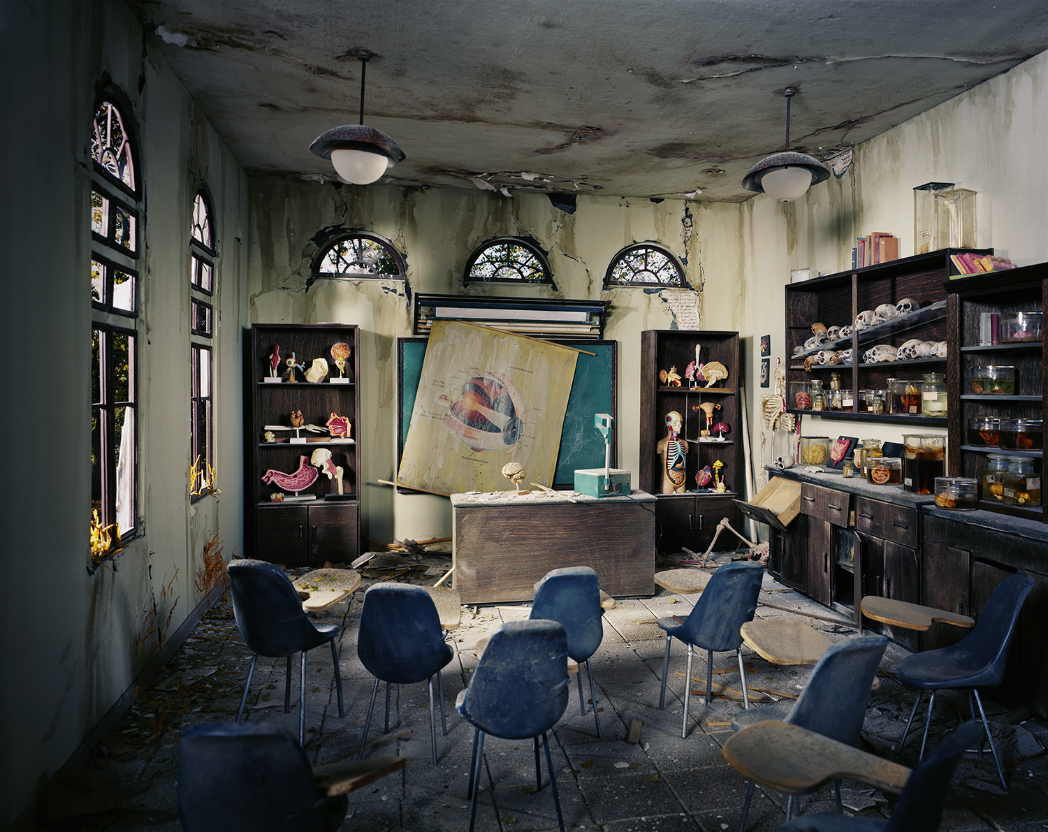 An abandoned classroom with decaying plaster walls and ceiling features modern blue chairs arranged around a wooden desk. Dark wood shelving units line the walls, displaying anatomical models, skulls, and educational materials beneath three arched windows. Anatomical diagrams and a skeleton figure are propped against the walls and desk, creating a haunting tableau of neglect and institutional decay.