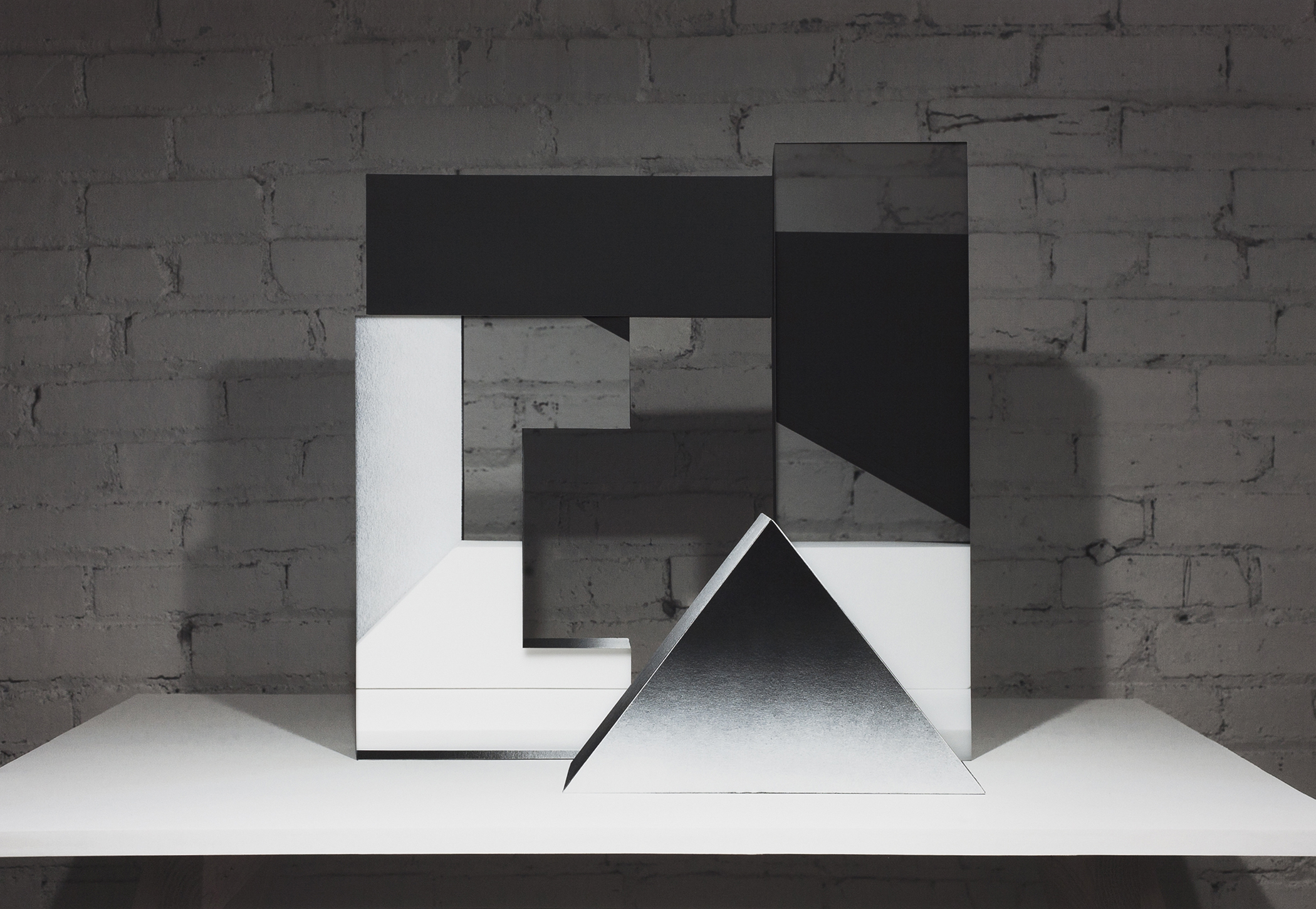 Abstract geometric composition featuring layered black and white rectangular forms arranged in an interlocking pattern against a weathered brick wall. A white triangular prism sits prominently in the foreground on a white platform, casting shadows that emphasize its three-dimensional form.