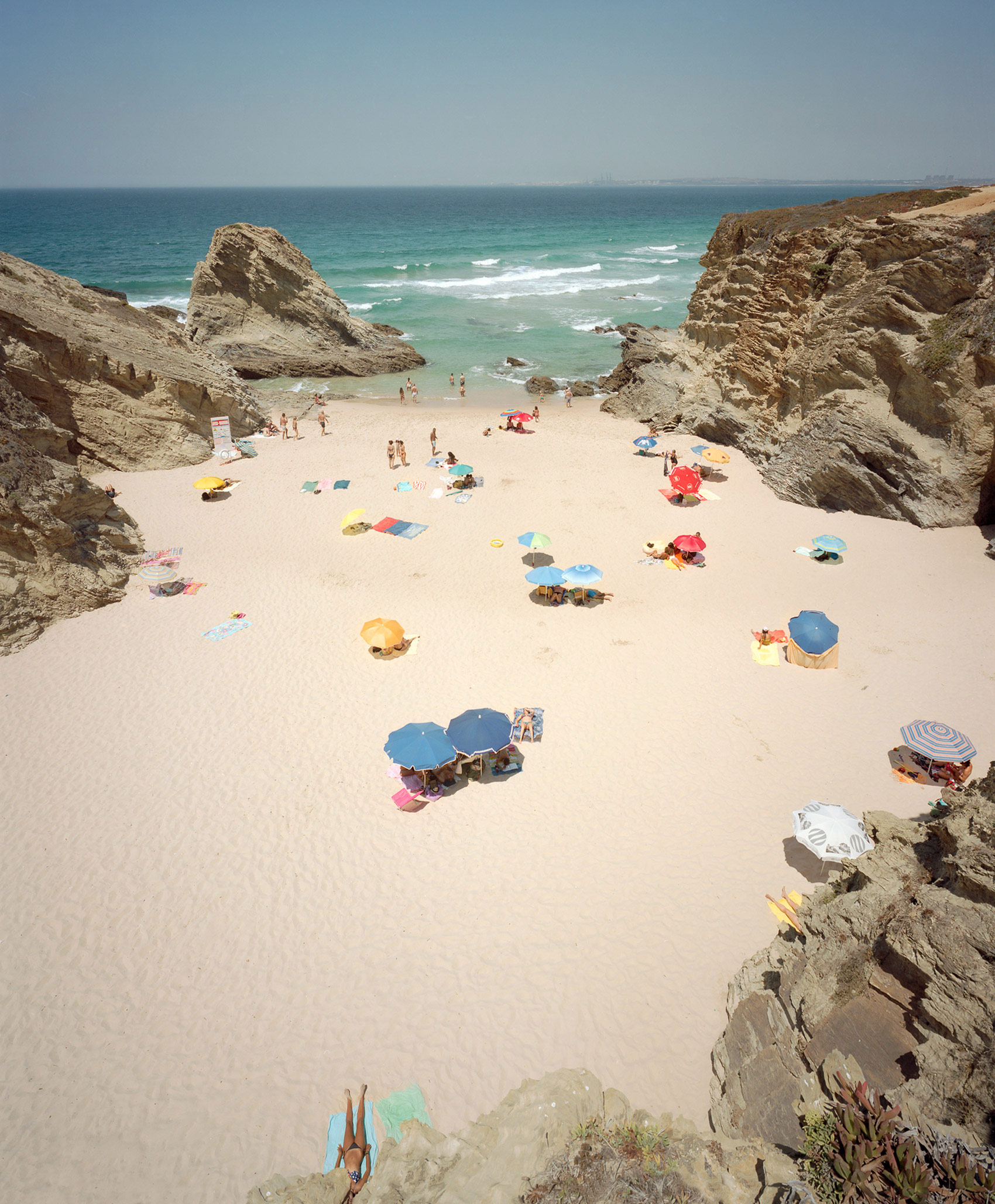 Aerial view of a secluded sandy cove bordered by dramatic limestone cliffs, with beachgoers scattered across the sand beneath colorful umbrellas and towels, turquoise waves visible beyond.