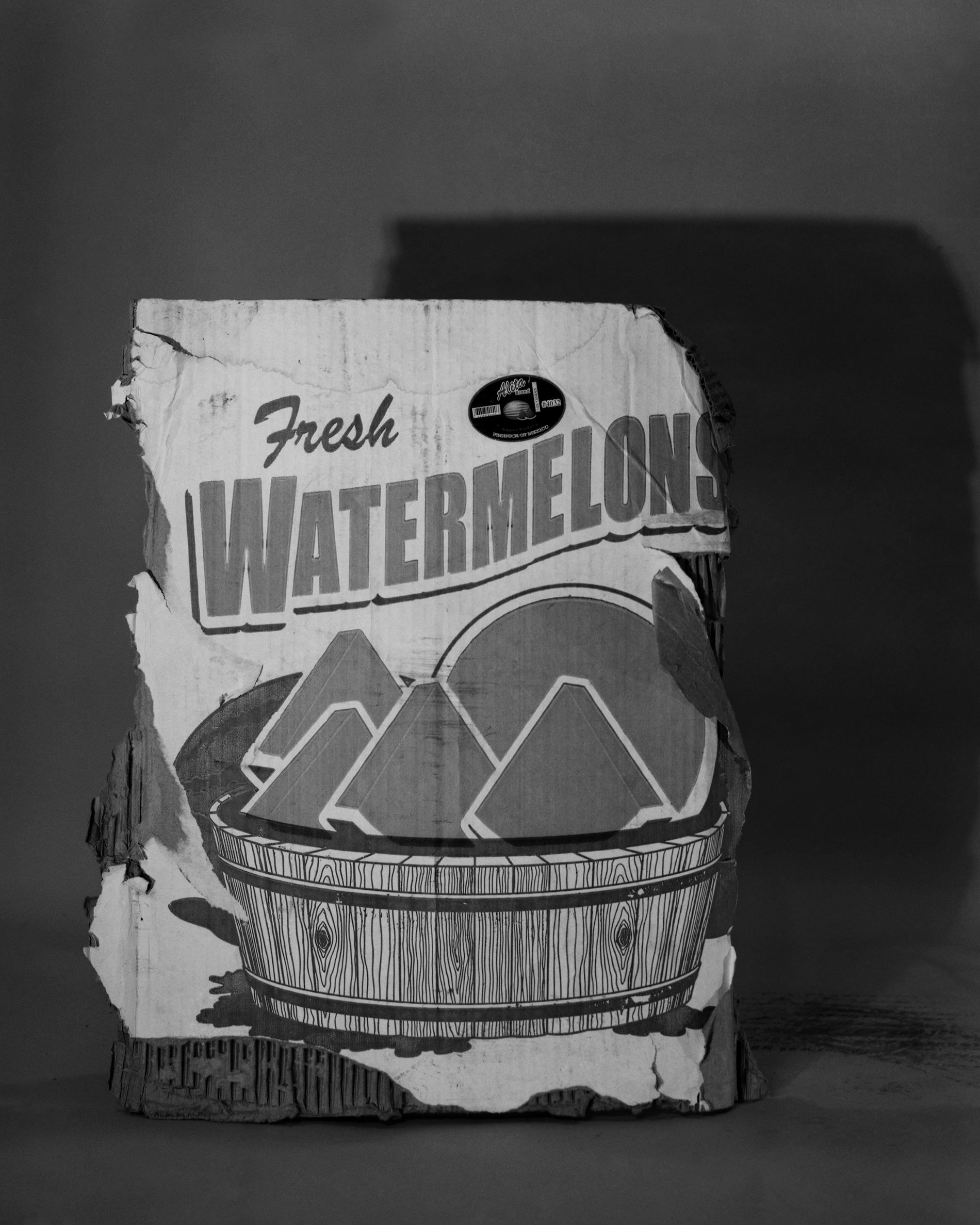A weathered vintage watermelon crate label sits against a neutral background in black and white. The label features bold typography reading "Fresh Watermelons" above an illustration of three stylized watermelon wedges nestled in a wooden barrel, rendered in a classic mid-century graphic style.