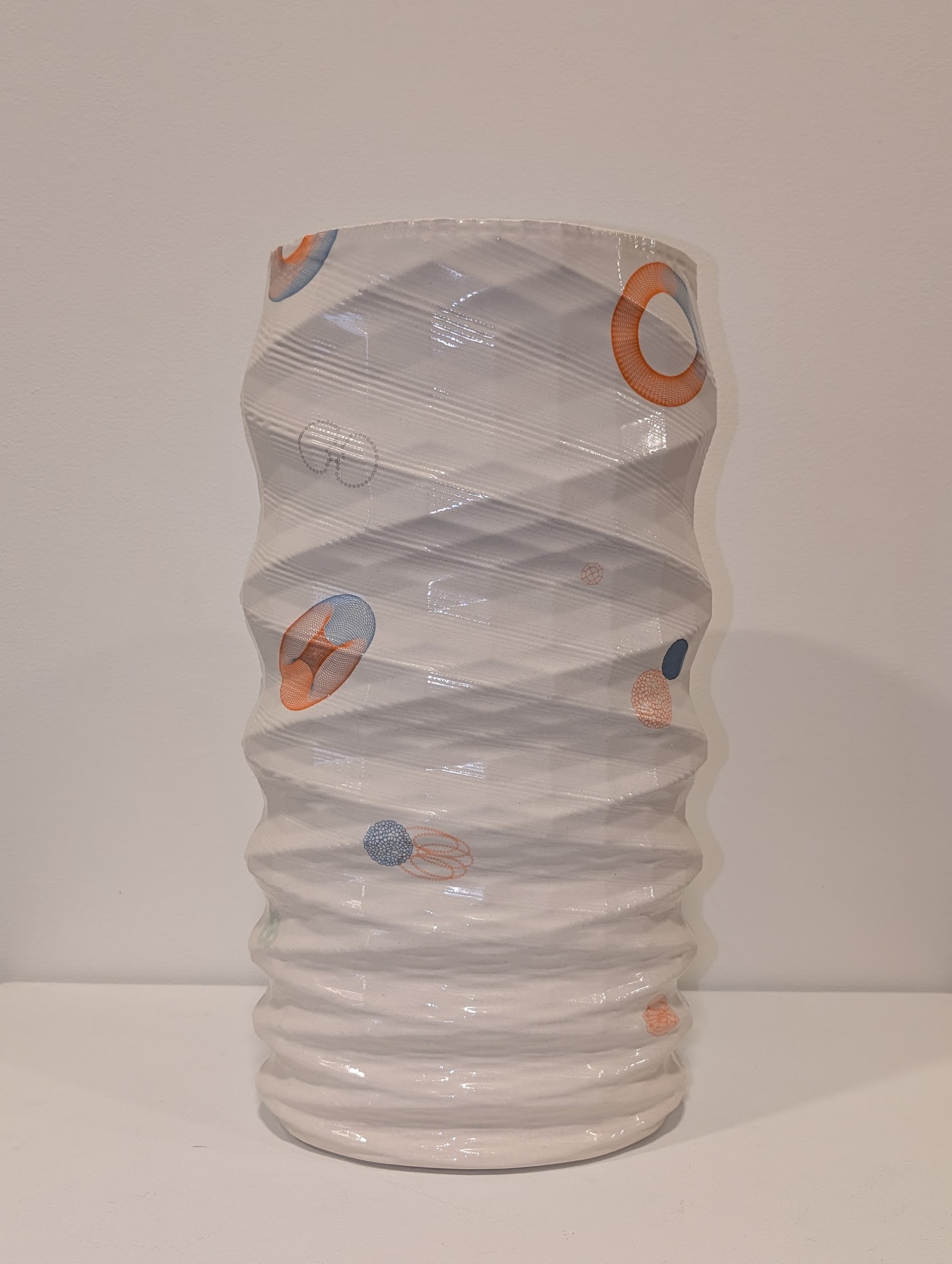 A translucent cylindrical vessel with horizontal ridged texture, wrapped in diagonal bands of clear material. Small abstract geometric shapes in orange, blue, and coral tones are scattered across the surface, creating a sense of movement around the form.