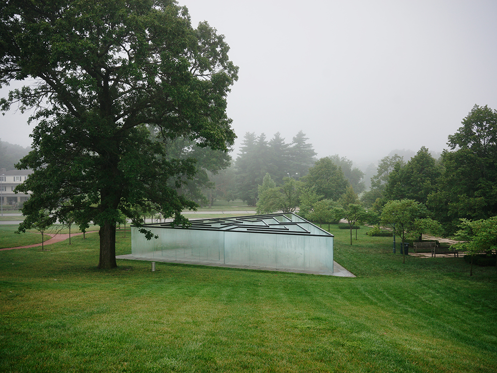 A contemporary white geometric structure with linear black roof elements sits on manicured green lawn beneath a large mature oak tree, surrounded by misty woodland and distant institutional buildings in soft overcast light.