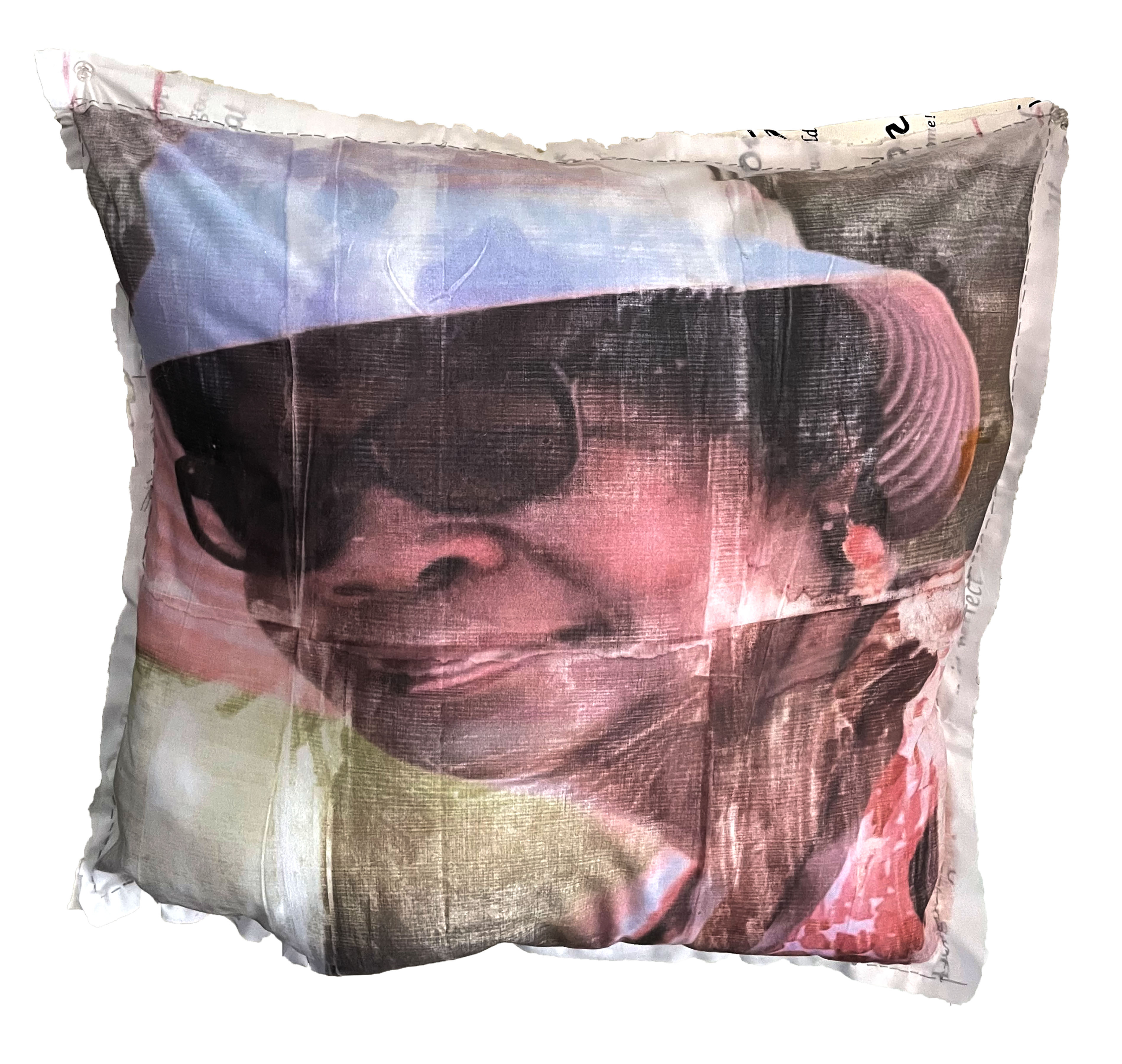 A translucent textile pillow features a portrait of a woman wearing sunglasses, rendered in soft pink, blue, and dark tones. The image appears layered and ethereal, with the figure's face and upper body visible through the semi-transparent fabric material, creating an atmospheric, dreamlike quality.