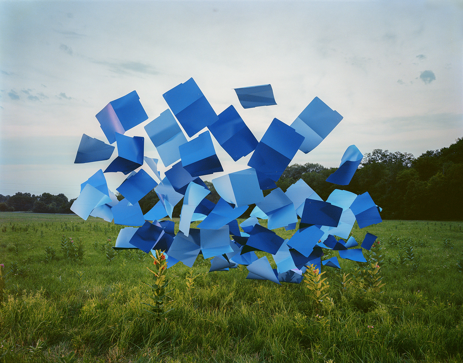 A large abstract sculpture composed of varying shades of blue rectangular and geometric forms stands in an open green field, with trees visible along the horizon under an overcast sky.