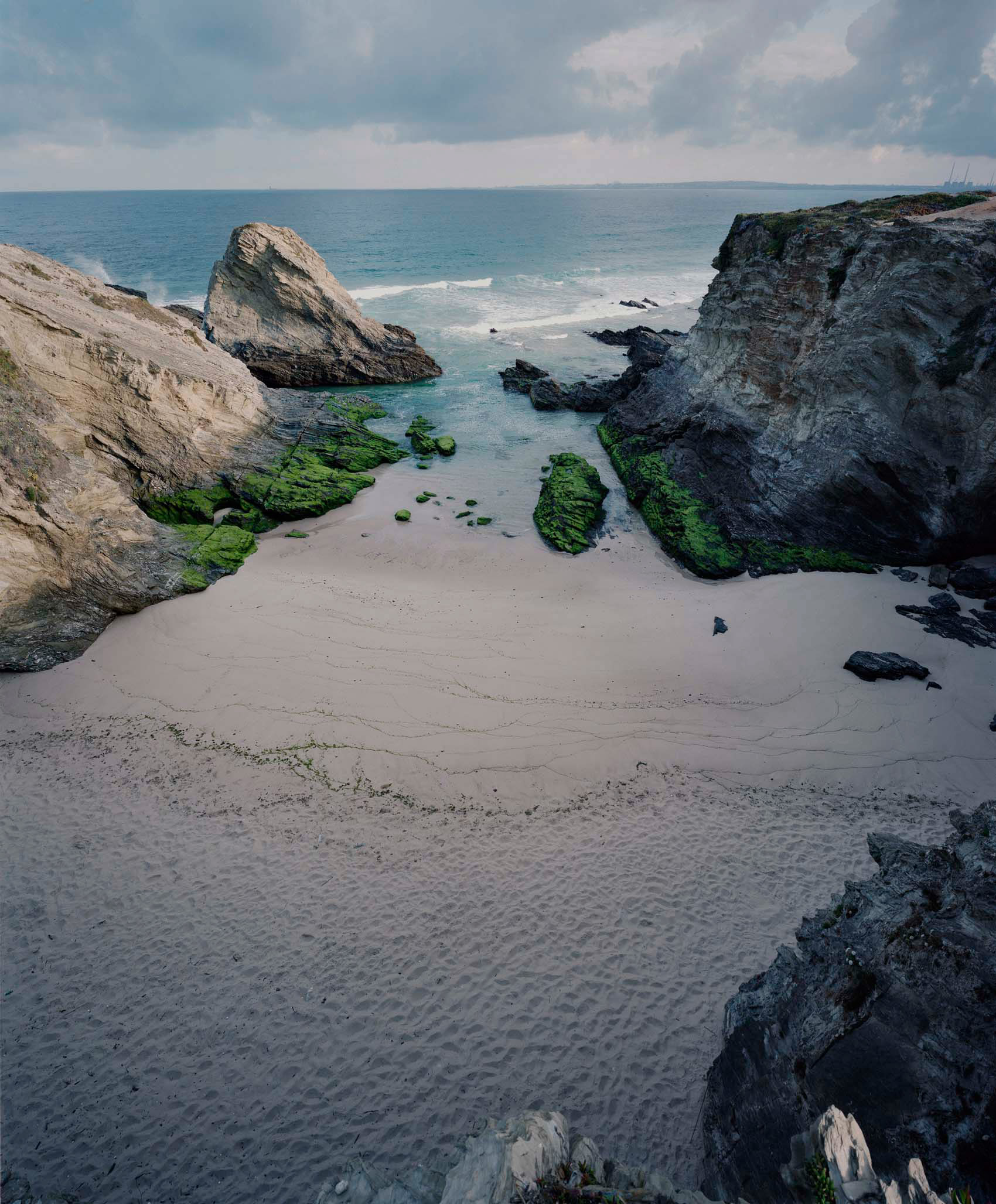 A rocky cove with pale sand nestled between layered gray and white cliff formations. Vibrant green algae-covered rocks emerge from shallow turquoise water, while the open sea stretches beyond under a clouded sky.