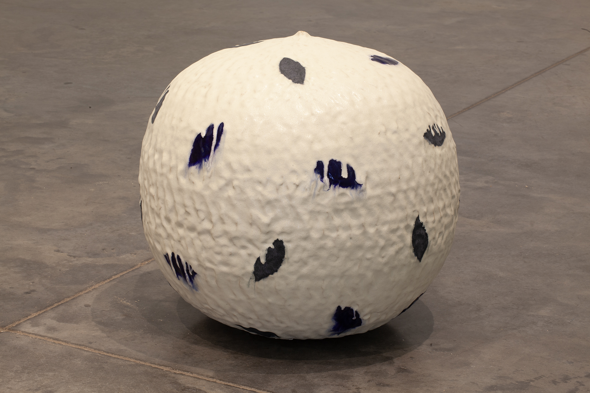 A large cream-colored spherical ceramic form sits on a concrete floor, its surface marked with gestural brushstrokes in deep blue and charcoal gray that resemble organic marks or leaf impressions distributed across the rounded form.