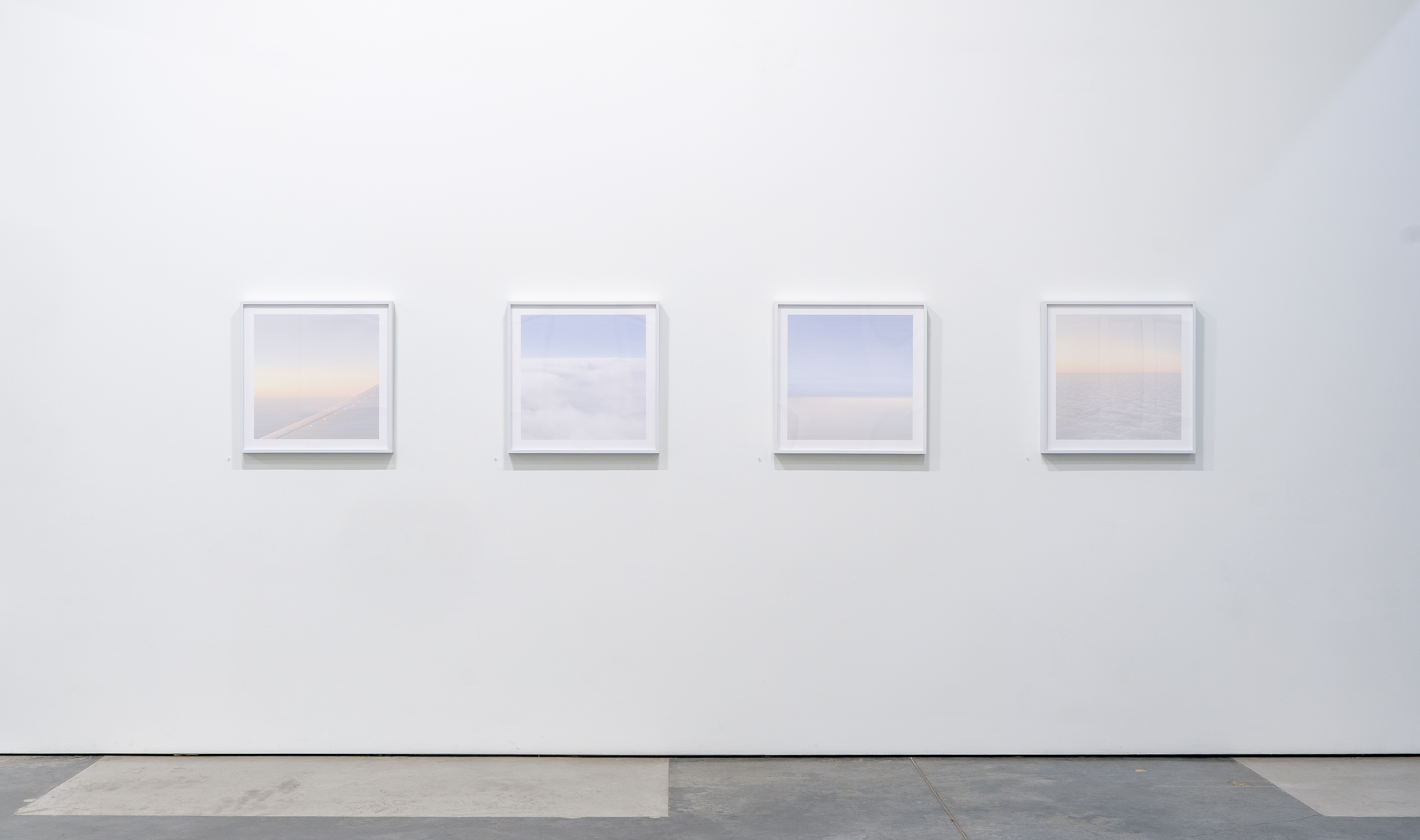 Four framed photographs arranged horizontally on a white gallery wall, each depicting serene aerial views of clouds and sky in soft blues, whites, and pale peachy tones, rendered in a subtle dotted texture pattern.