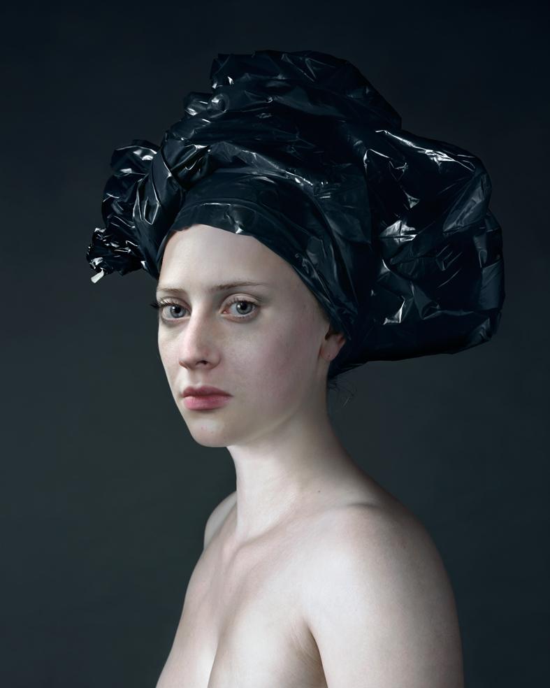 Portrait of a pale-skinned person with a neutral expression, shown in three-quarter profile against a dark gray background. Their head is wrapped in crumpled black plastic material that extends upward and outward, creating an elaborate sculptural headdress. The figure's shoulders are bare, emphasizing the stark contrast between skin tone and the glossy black material.