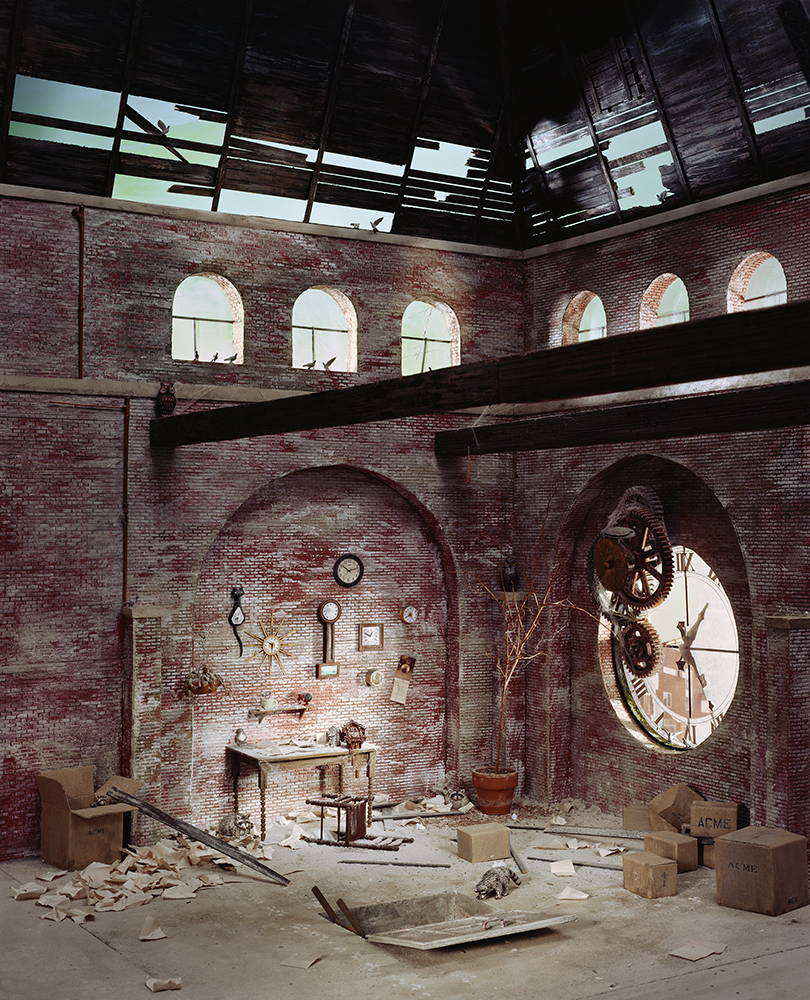 An abandoned brick interior with a partially collapsed ceiling reveals sky through broken wooden beams. A large gear-driven clock face protrudes from the right wall, while the left alcove displays hanging clocks and mechanical objects amid scattered debris and cardboard boxes labeled "ACME" on the floor below.