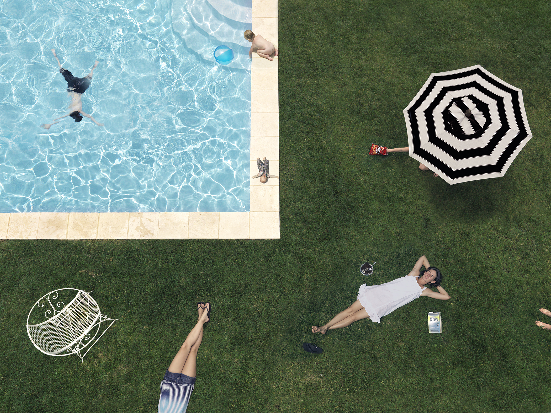 Aerial view of a backyard divided between a swimming pool and lawn, where multiple figures engage in leisure activities. Two swimmers float in bright turquoise water while a person stands poolside; on the grass below, individuals recline under striped umbrellas, with personal belongings scattered nearby.