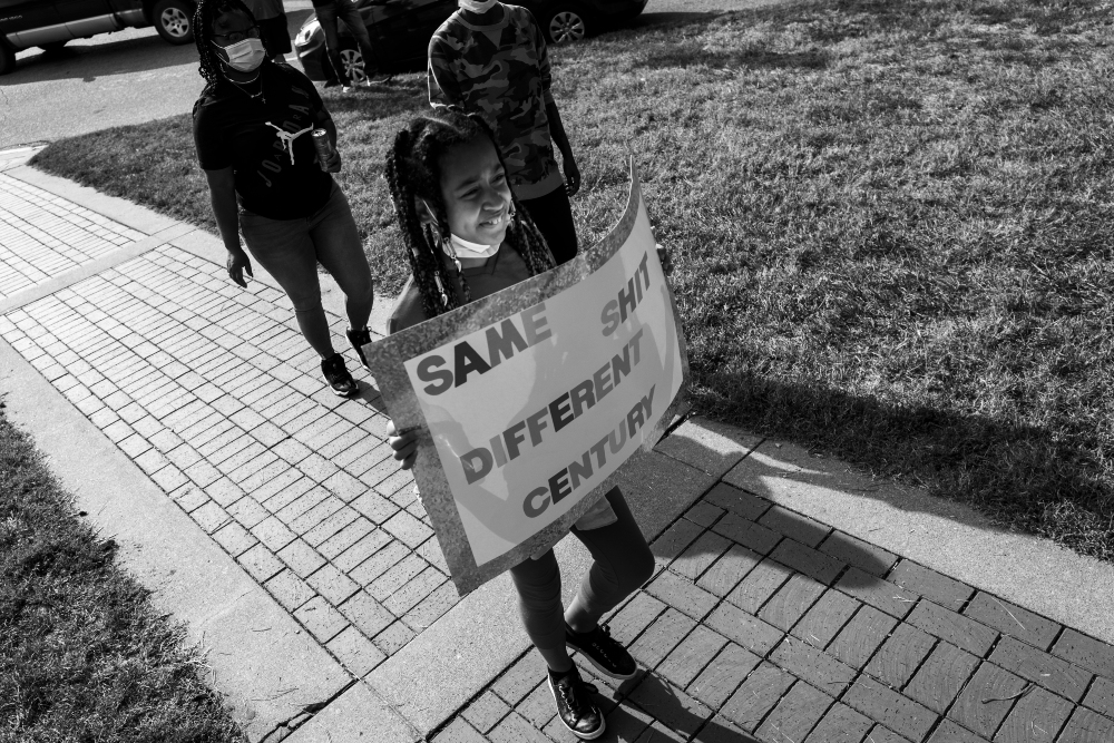 Black and white photograph of activists on a sidewalk holding a protest banner that reads "SAME SHIT DIFFERENT CENTURY." Multiple figures wearing masks stand together on a paved path bordered by grass, with parked cars visible in the background.