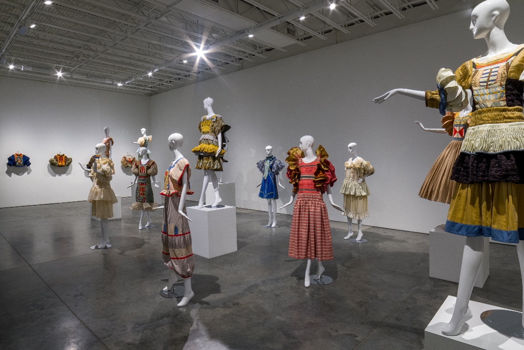 A gallery installation featuring mannequins dressed in elaborately constructed garments with vibrant colors including golds, reds, blues, and creams. The garments showcase intricate hand-pleating, embroidery, and patchwork details, displayed on white pedestals throughout a minimalist white-walled space with polished concrete floors. Two small fabric pieces hang on the left wall. The clothing ranges from sculptural silhouettes with voluminous skirts to fitted bodices, blending historical costume references with contemporary fiber art techniques.