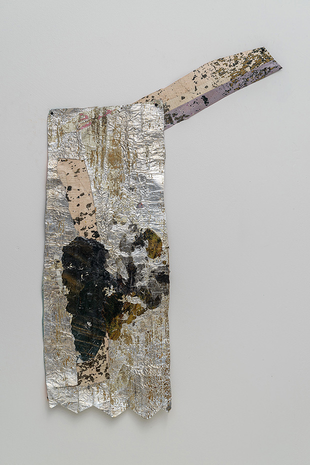 A textured mixed-media artwork composed of crumpled and layered metallic foil in silver and gold tones, featuring areas of black charring or burn marks and tan transfer-printed elements. The piece is mounted at an angle against a white wall, with a rectangular main section and an extended arm-like extension decorated with scattered dark speckles and layered materials.