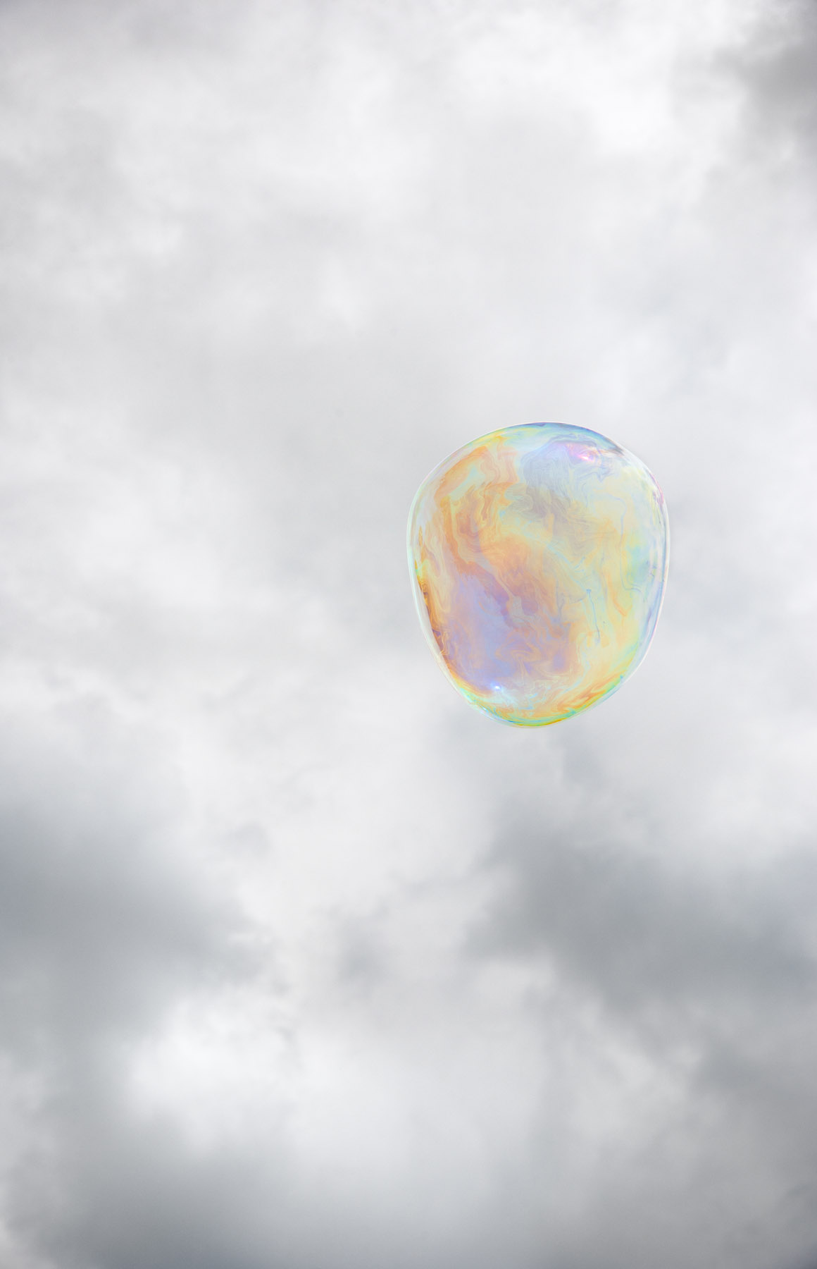A translucent soap bubble floats against a soft, cloudy gray sky. Its spherical surface displays iridescent swirls of pastel yellow, pink, blue, and green that shift across the membrane.