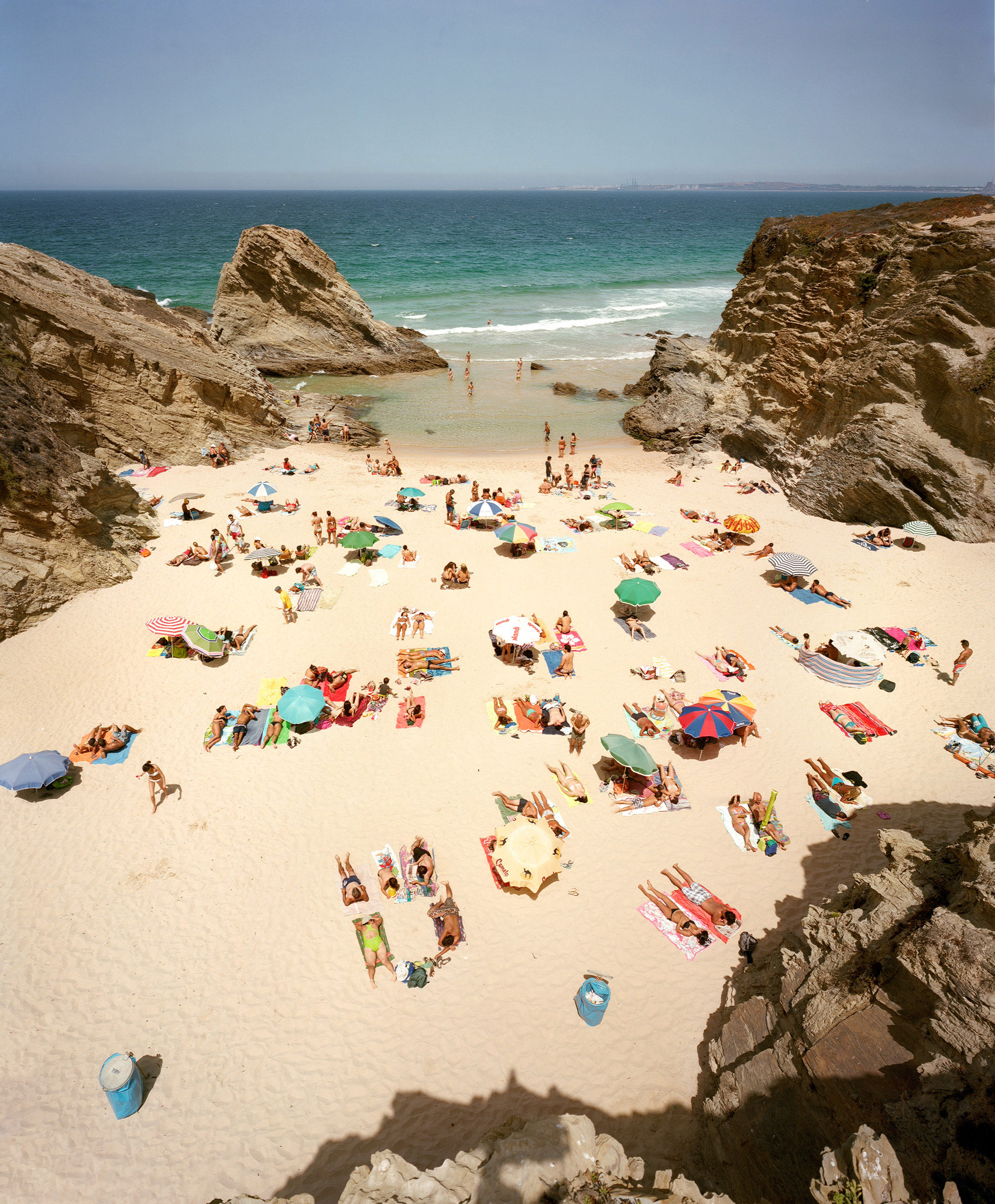 Aerial view of a crowded beach cove framed by dramatic rocky outcrops, with dozens of beachgoers scattered across pale sand beneath colorful umbrellas and towels, turquoise water visible beyond the shore.
