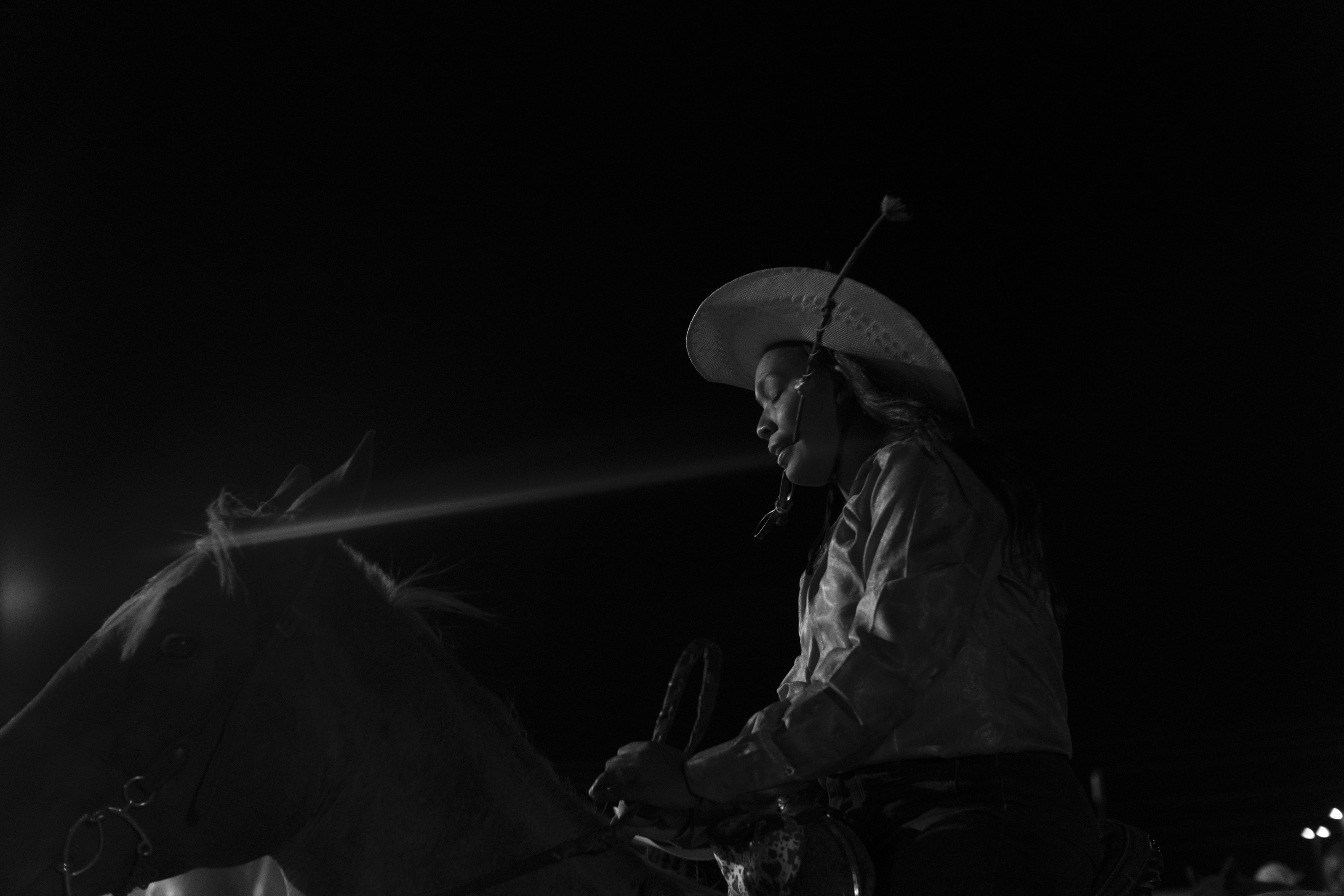 Black and white photograph of a figure wearing a wide-brimmed hat and long coat, seated on a horse against a dark background. The composition is rendered in high contrast, with dramatic lighting that emphasizes the silhouettes of both rider and horse.