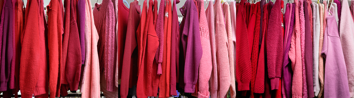 Multiple red and pink sweaters and cardigans of varying shades hang closely together on a rack, their textured knit fabrics creating a dense, monochromatic composition in warm tones ranging from deep crimson to pale rose.