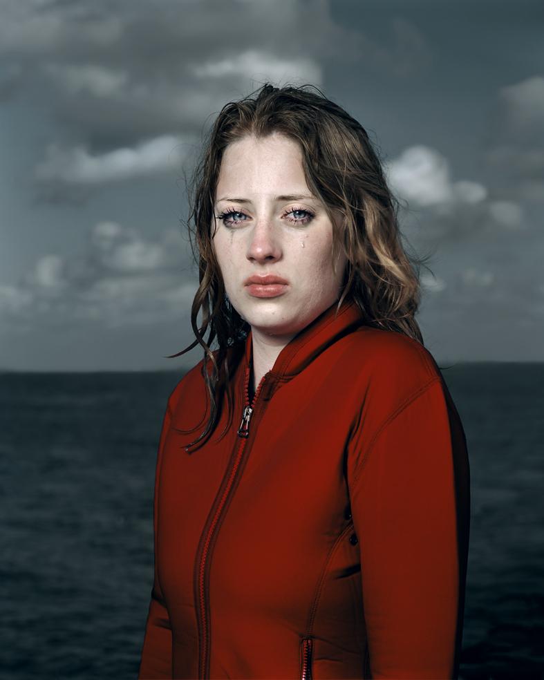 Portrait of a woman with dark, wet-looking hair wearing a bright red zip-up jacket, photographed against a stormy seascape with overcast sky and turbulent water.