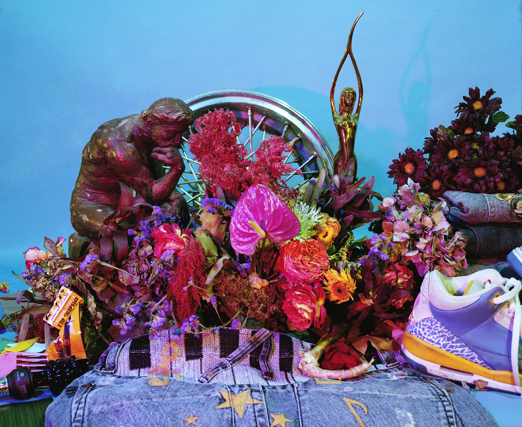 A vibrant still life composition against a bright blue background featuring an ornate bronze sculpture of a figure on the left, surrounded by an abundant arrangement of magenta, pink, and deep purple flowers including roses and chrysanthemums. A golden figurative sculpture rises from the center, while everyday objects—a purple and yellow sneaker, denim fabric with star patterns, striped textile, and various personal items—are integrated throughout the floral display, creating a layered collision of fine art, nature, and consumer culture.
