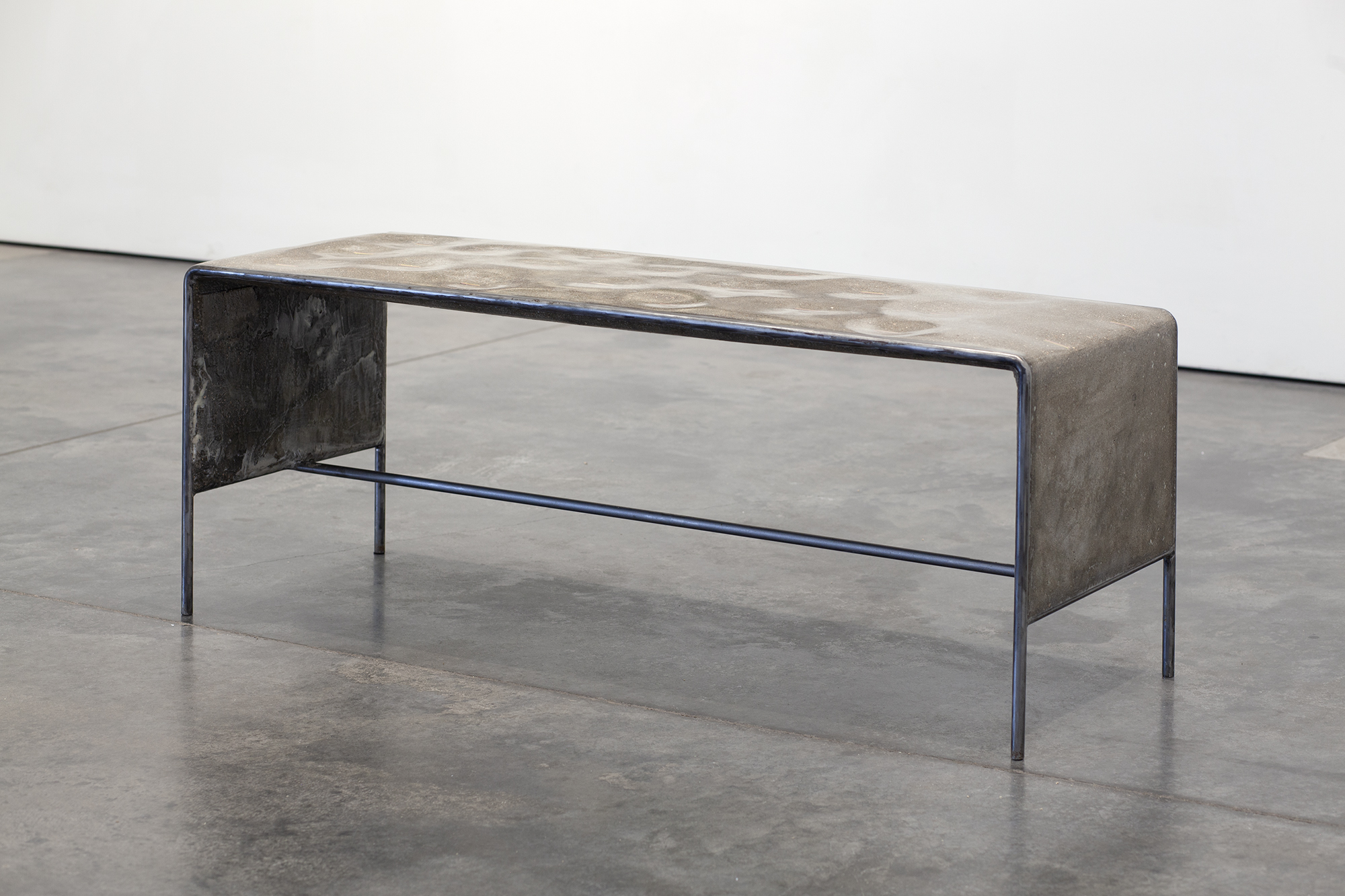 Industrial bench with a concrete and ceramic top featuring geometric surface patterns, supported by dark metal legs and frame with blue-tinted steel accents. The weathered materials and minimalist construction create a stark, contemporary sculptural form displayed on polished concrete flooring.