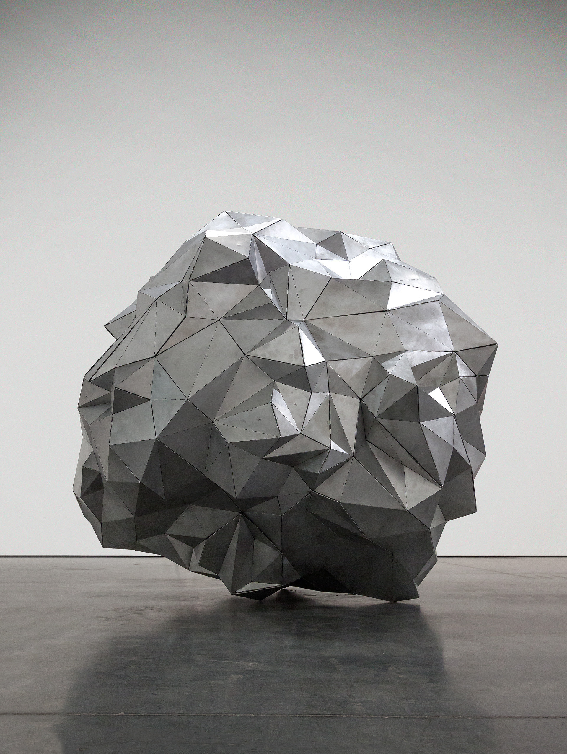 A large spherical sculpture composed of folded aluminum planes creates a faceted, geometric surface. The form is rendered in shades of gray and white, with dramatic shadows cast by the angular facets, set against a neutral gallery wall and polished concrete floor.