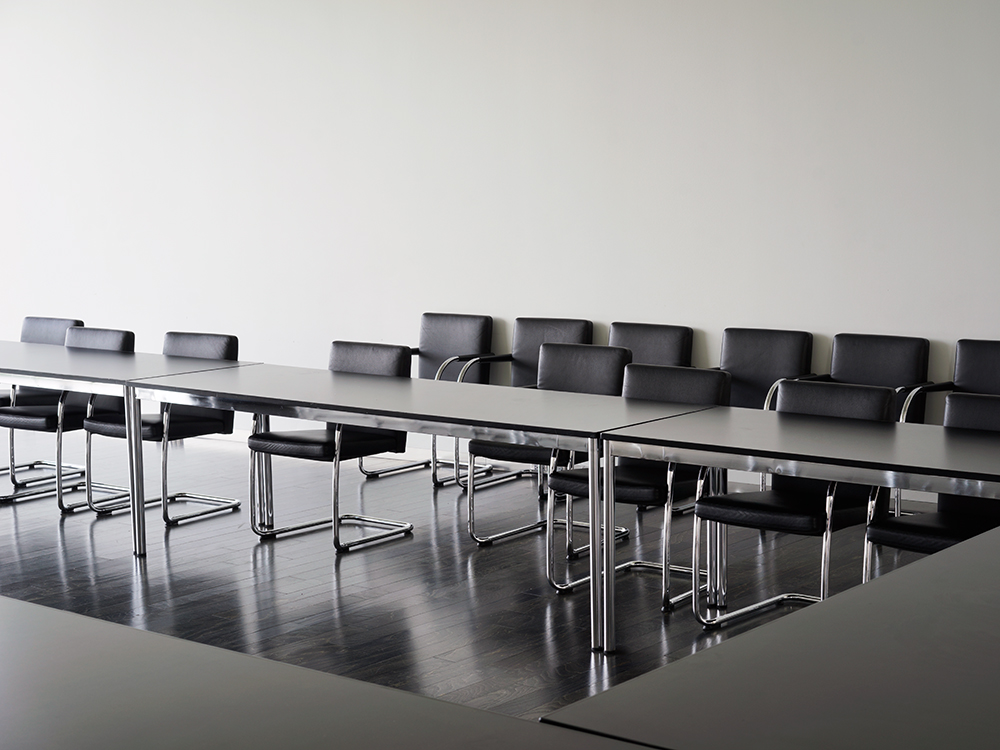 A minimalist interior photograph depicting a sleek conference room with a long, reflective dark table surrounded by black leather chairs with chrome frames. The polished dark flooring mirrors the table's surface, while white walls and subtle overhead lighting create a stark, contemporary aesthetic emphasizing geometric forms and material contrasts.