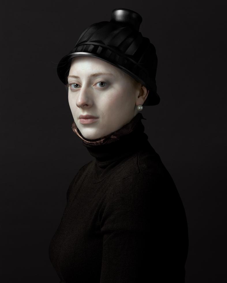 Portrait of a young woman wearing a black turtleneck and ornate black hat with decorative band, set against a dark background. She gazes toward the camera with a composed expression, her pale complexion and pearl earring subtly illuminated by controlled lighting that emphasizes the painting's classical Renaissance-inspired style.