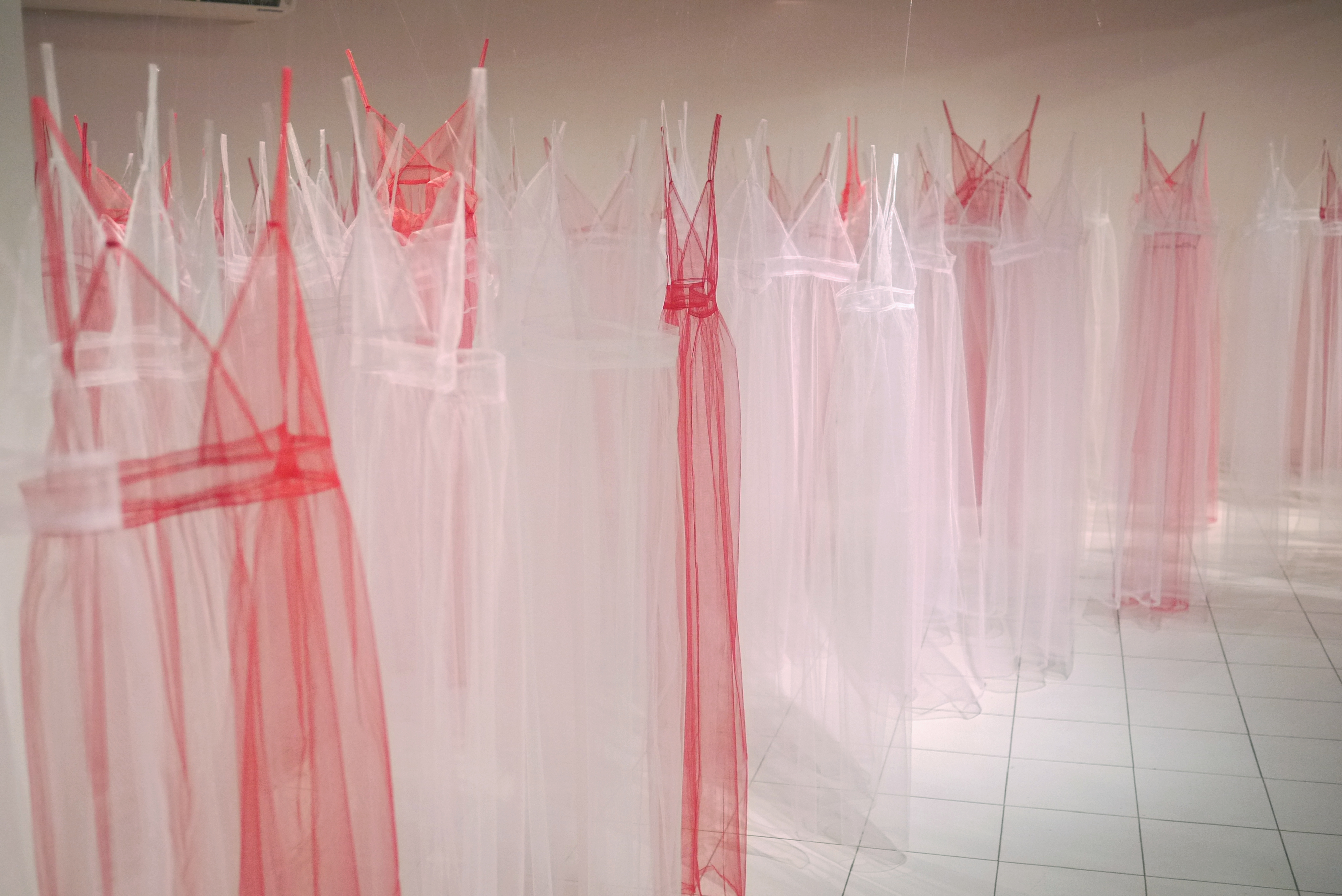 Multiple translucent tulle garments in shades of pink and white hang from above in a gallery space, their delicate fabrics creating layered, ethereal forms. The diaphanous dresses with extended straps cast soft shadows across the white tiled floor, establishing an installation that blurs the boundary between sculpture and fashion.