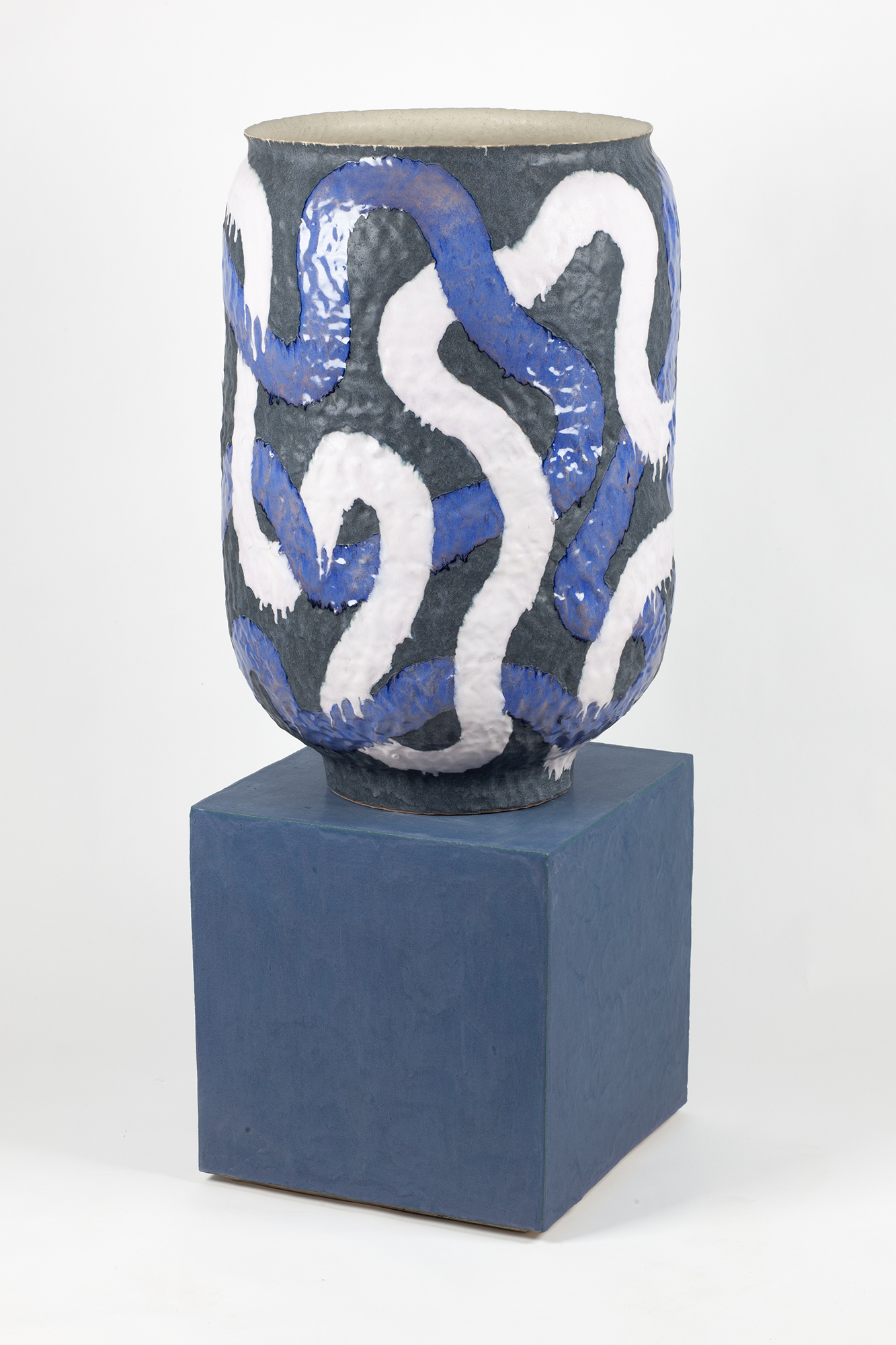 A cylindrical ceramic vessel with a cream-colored rim sits atop a blue cubic base. The vessel's dark gray body is decorated with intertwining blue and white vine-like forms that spiral around its surface, creating an organic, flowing pattern against the muted background.