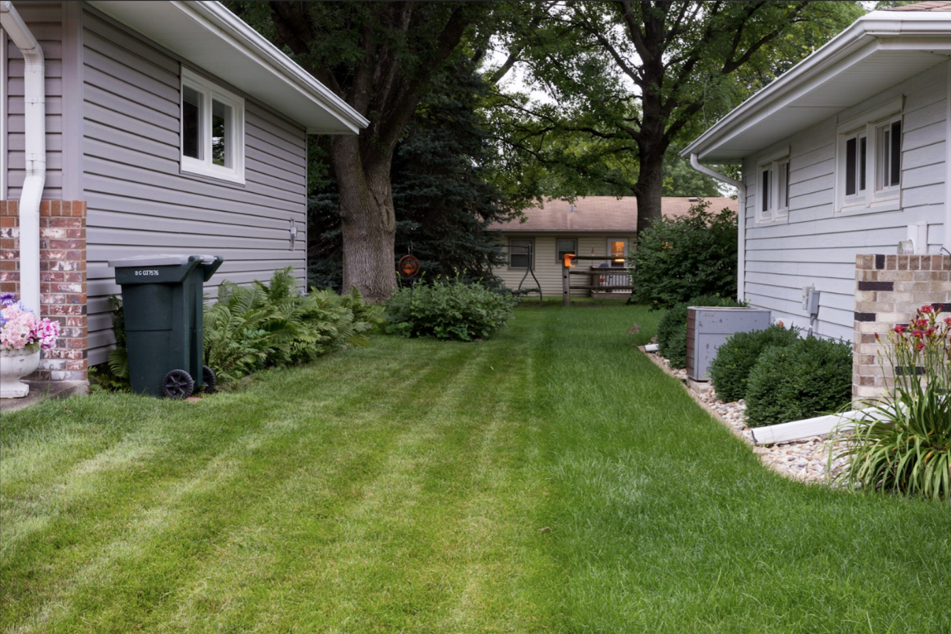 A residential backyard bordered by two houses with gray siding and brick chimneys, featuring a well-maintained lawn, mature trees, flowering plants along the edges, a trash bin, and utility boxes, rendered in vibrant greens and natural daylight.