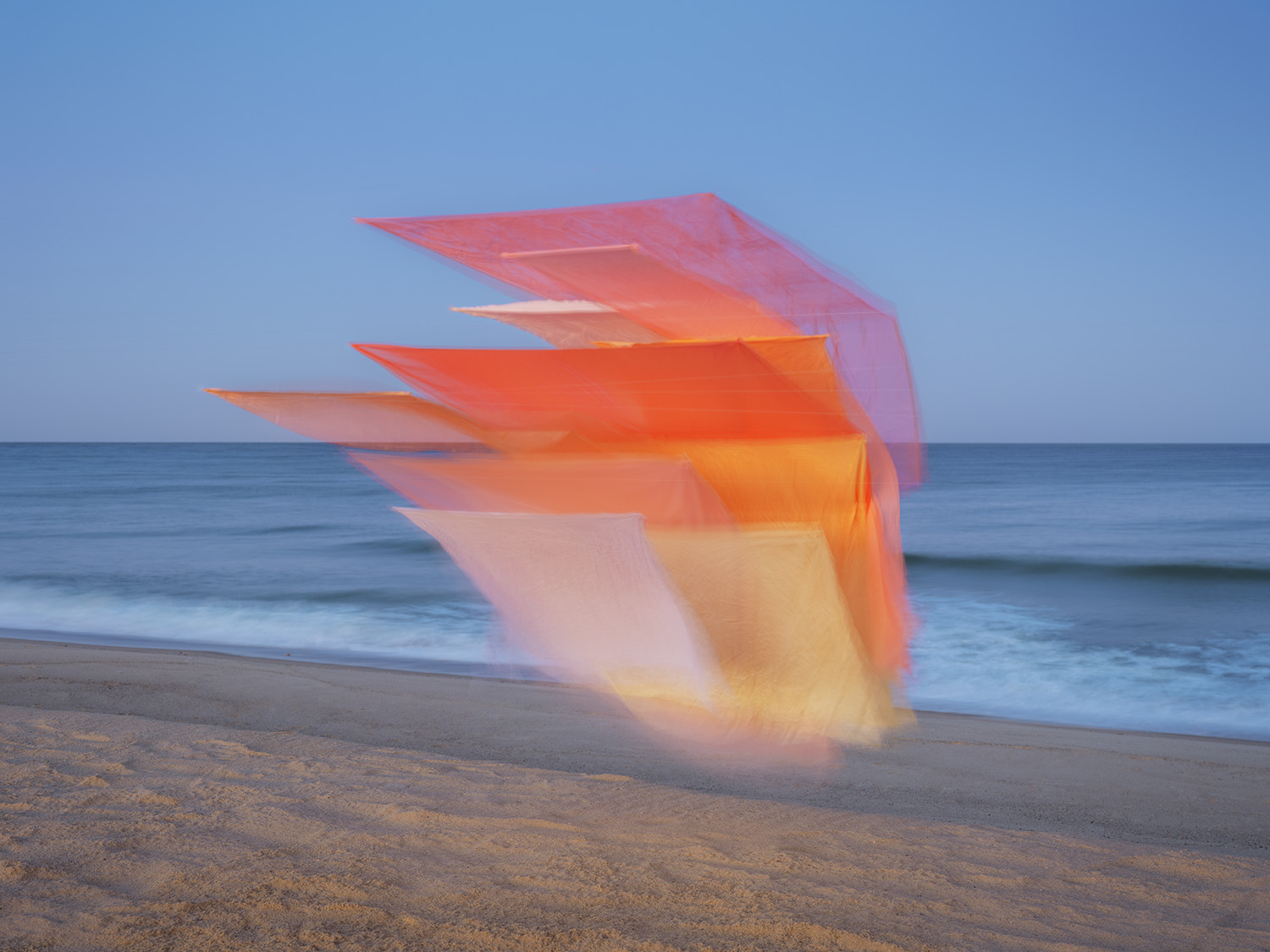Layered sheets of translucent silk in gradating shades of pink, coral, orange, and yellow billow across a beach, creating an ethereal geometric composition against the ocean and sky. The flowing fabric catches light and casts soft shadows on the sand, blending the vibrant colors in areas of overlap.